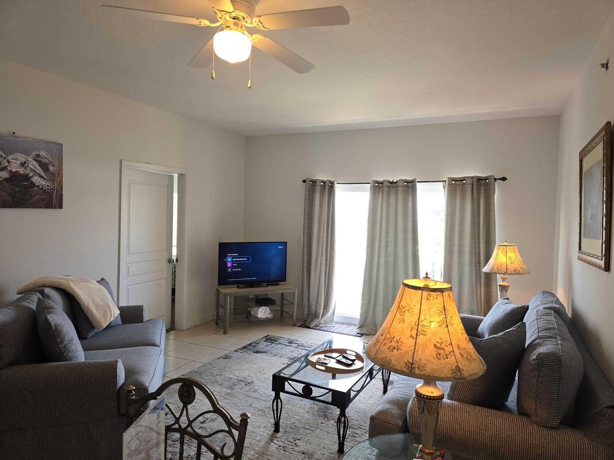 A living room with a ceiling fan, a television on a stand, a sofa, a coffee table, and a lamp.