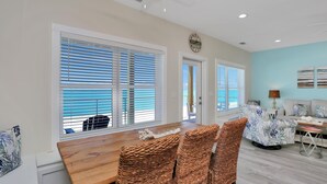 A dining area with a wooden table and wicker chairs, large windows with blinds, a sofa, and a lamp. - Beach Fantasies B (Port St. Joe)