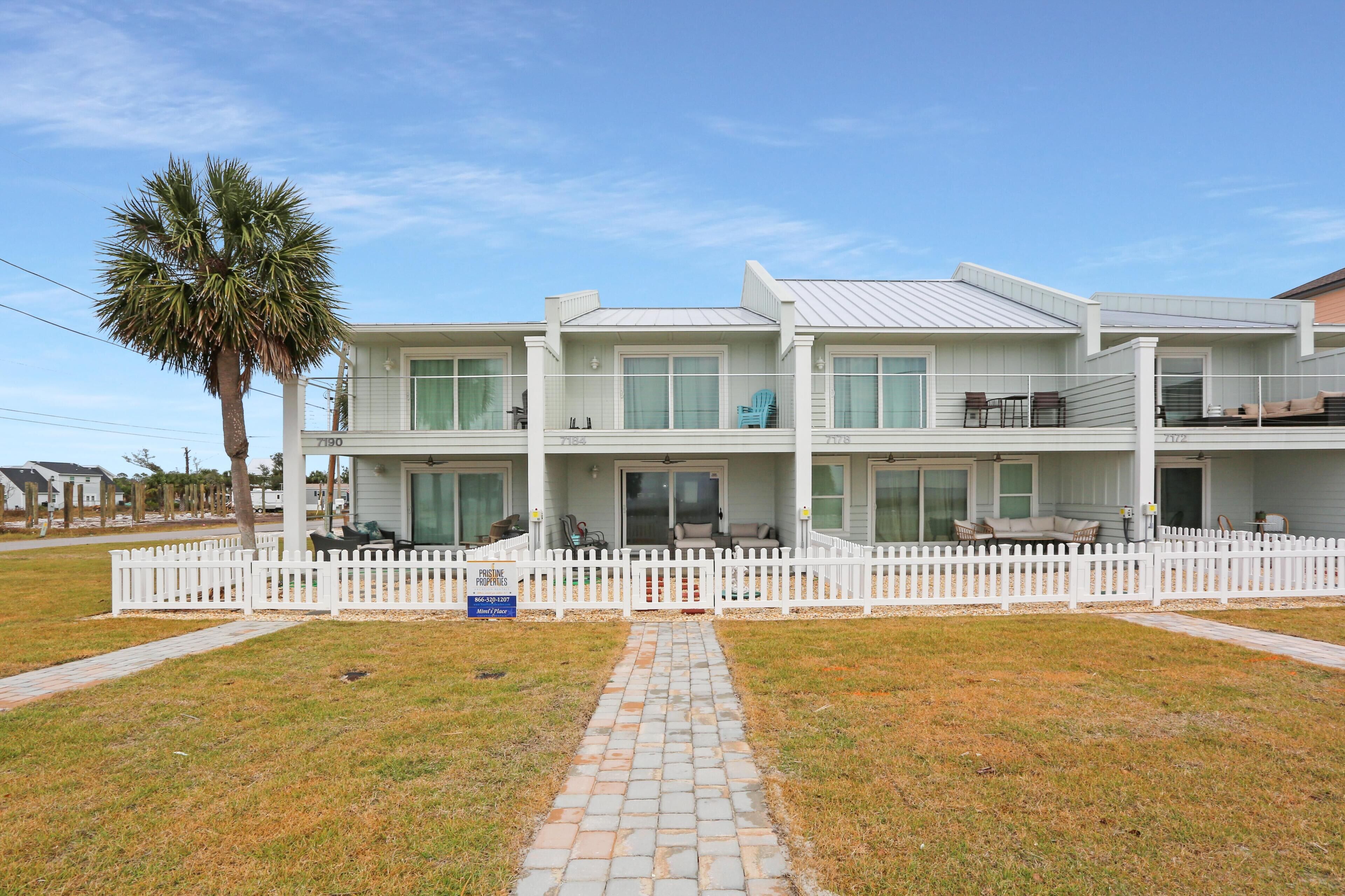 A two-story house with a white picket fence, a paved driveway, and a palm tree.