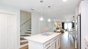 Une cuisine avec des meubles blancs, un îlot central, des luminaires suspendus et un escalier menant à un autre niveau.