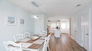 Un coin repas avec une table en bois et des chaises blanches, une cuisine en arrière-plan et un couloir menant à une porte.