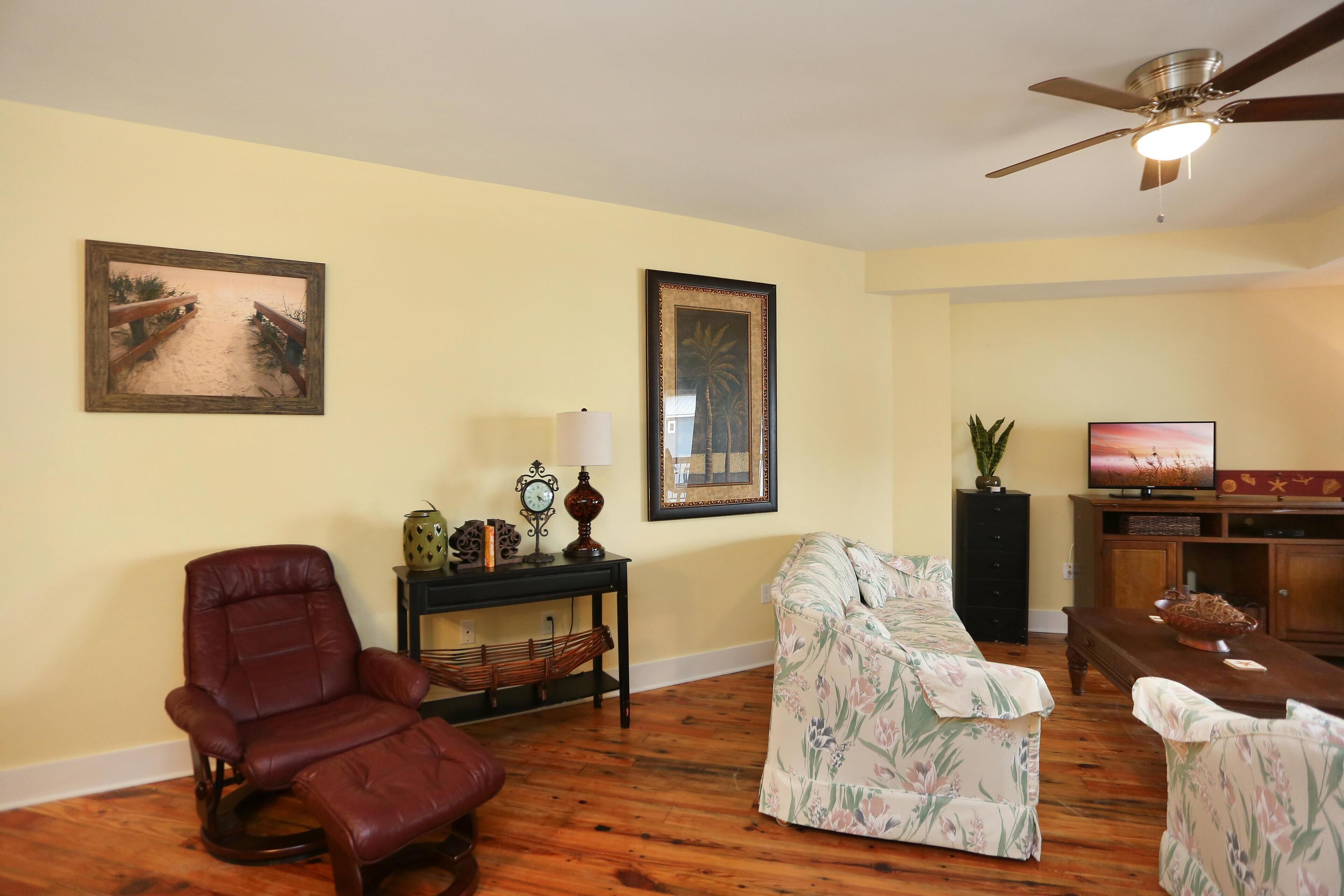 A living room with a brown leather chair, floral sofa, wooden floor, ceiling fan, and a television on a wooden stand.