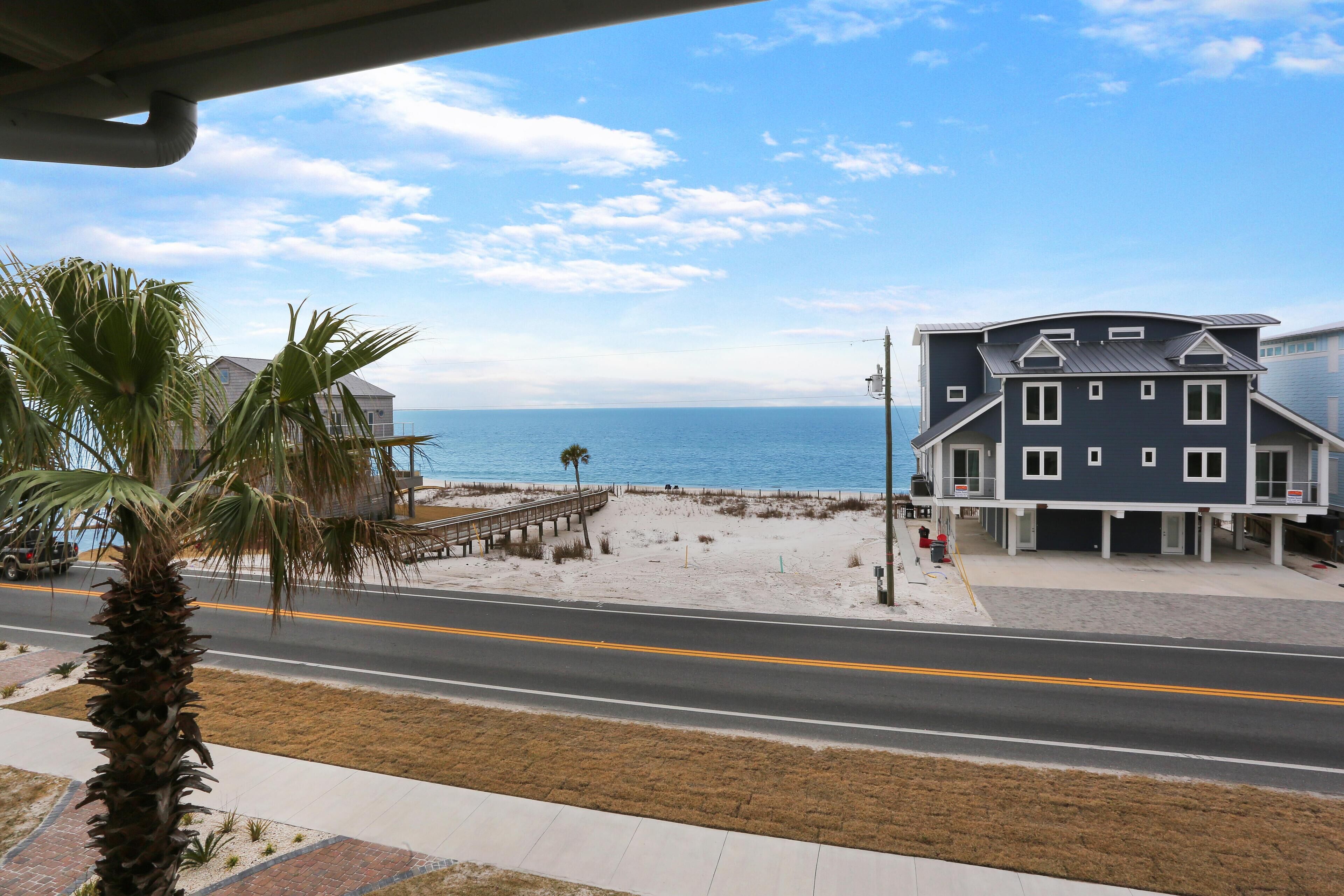 A coastal road with a beach, a blue building with a balcony, and a palm tree.