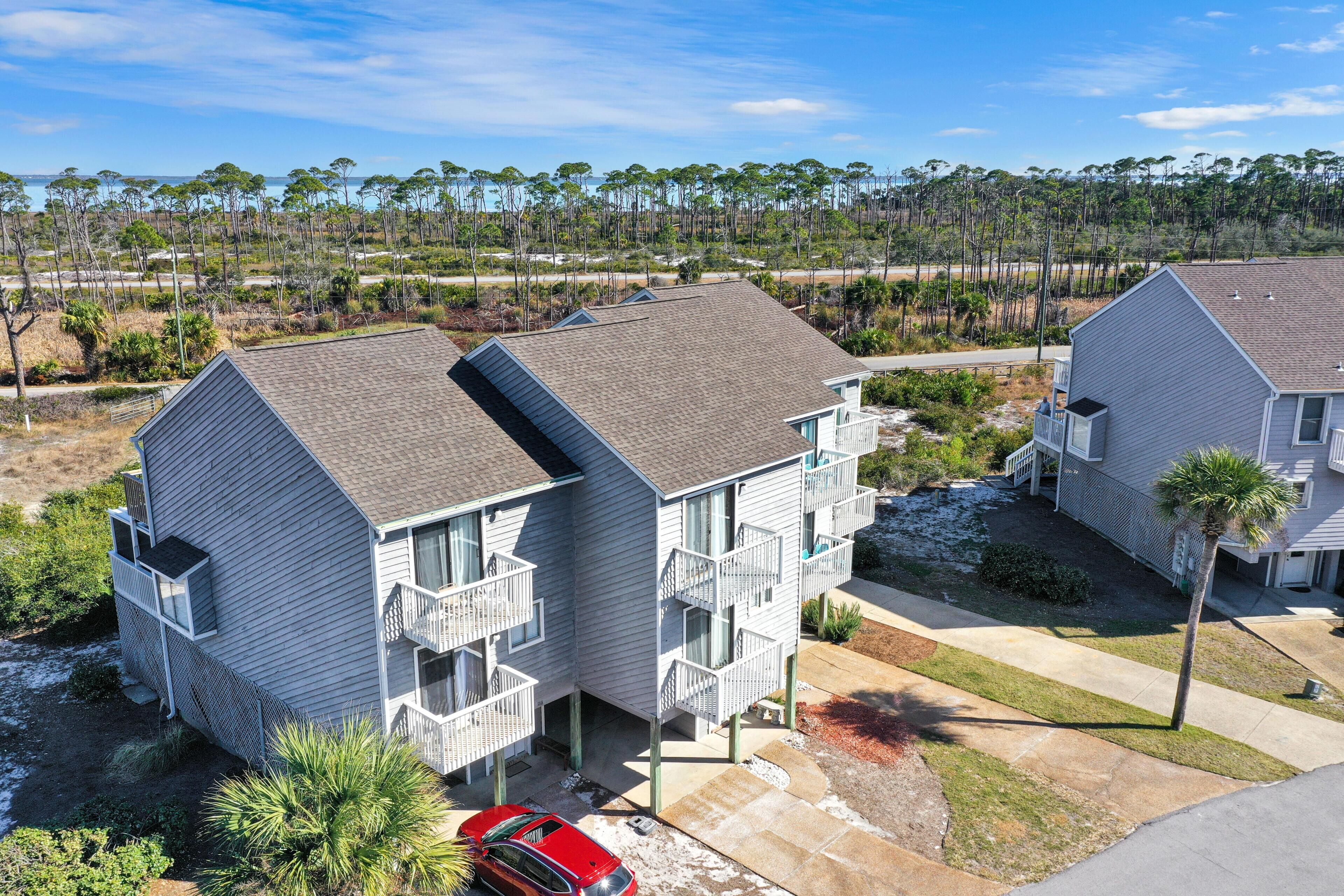 Um edifício residencial de dois andares com uma varanda, um carro vermelho estacionado na frente e uma vista para a praia e para as árvores ao fundo.