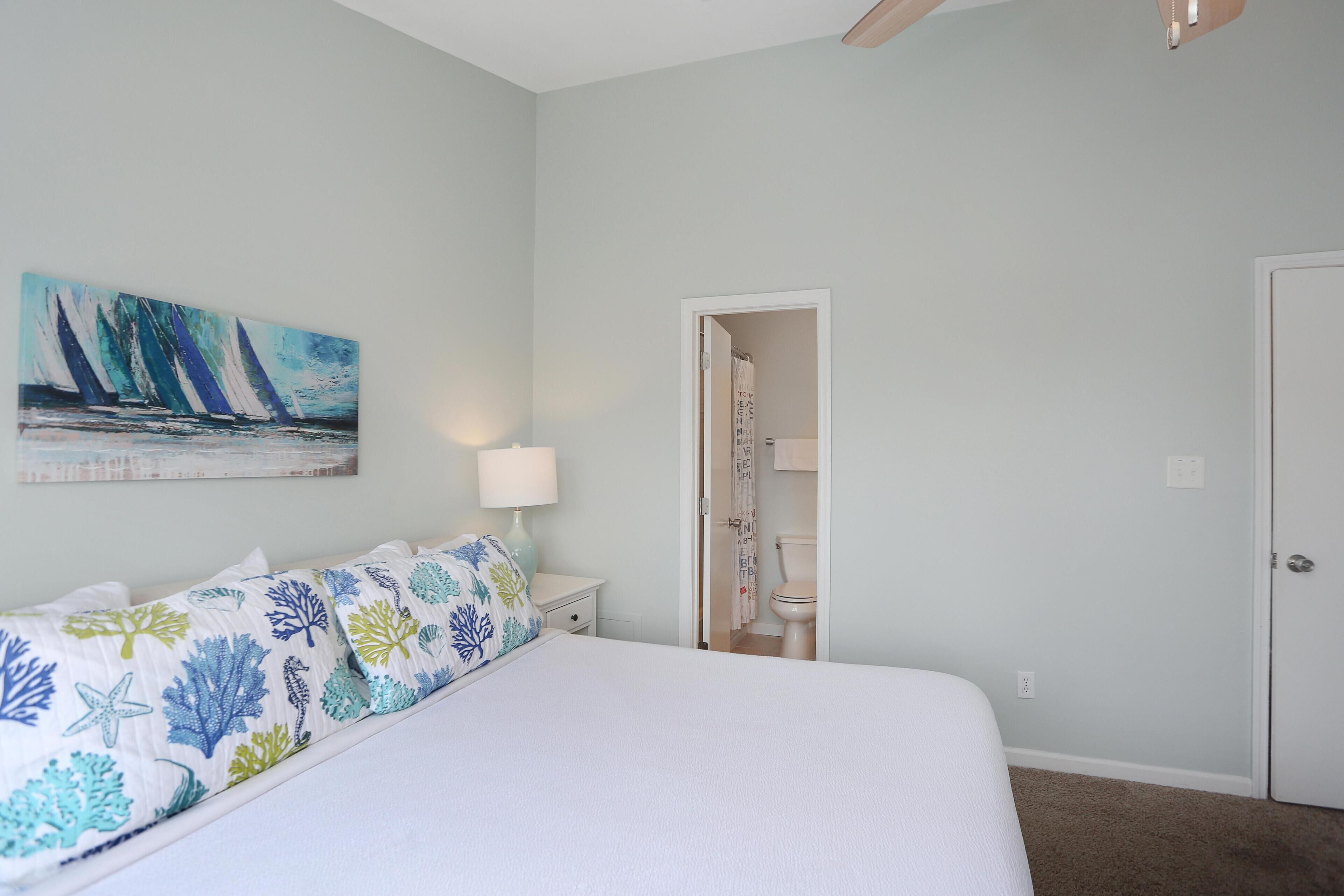 A bedroom with a bed, a nightstand, a lamp, a painting, and a door leading to a bathroom.