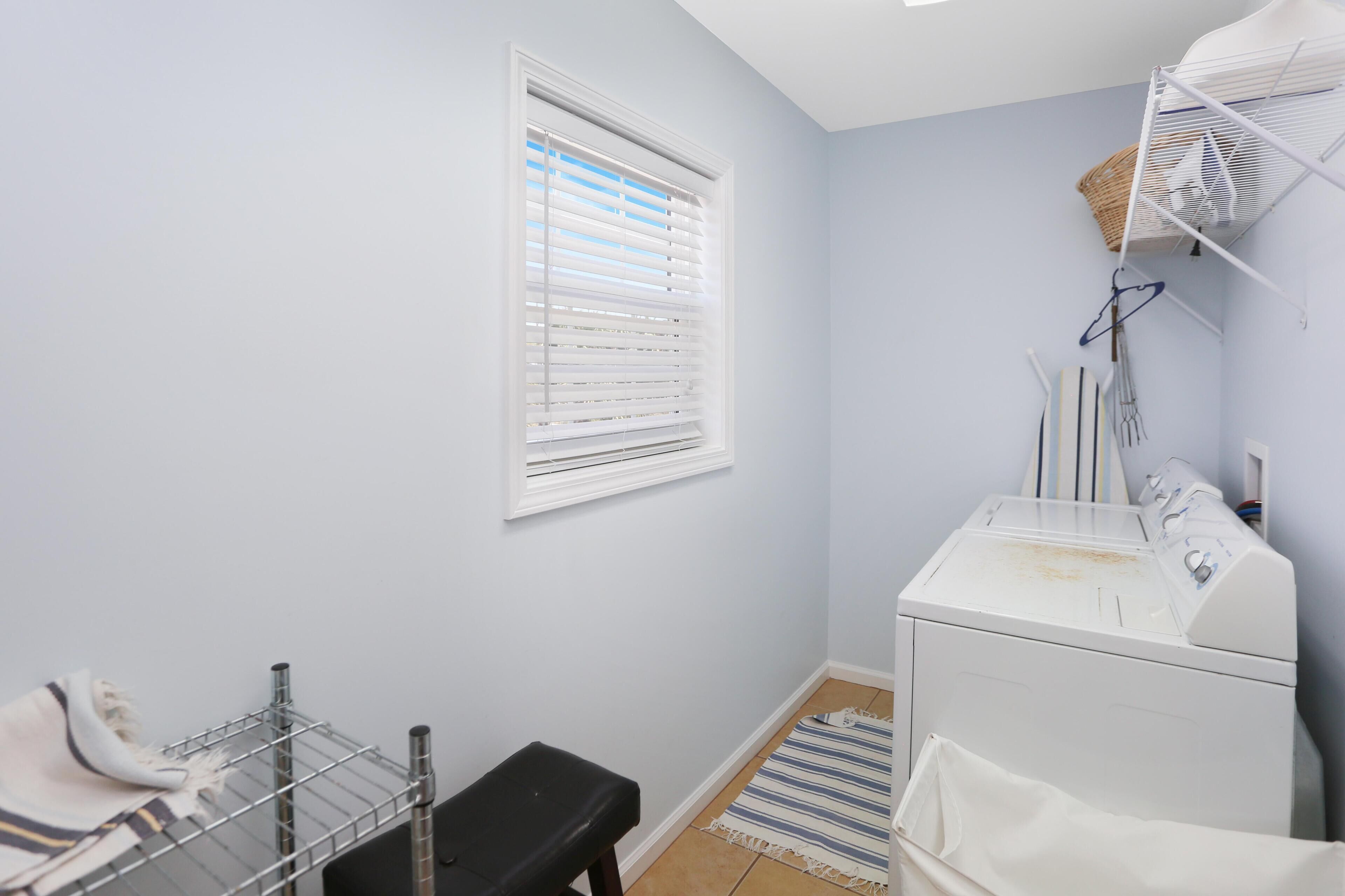 A laundry room with a washing machine, a drying rack, a bench, a window with blinds, and a striped rug.