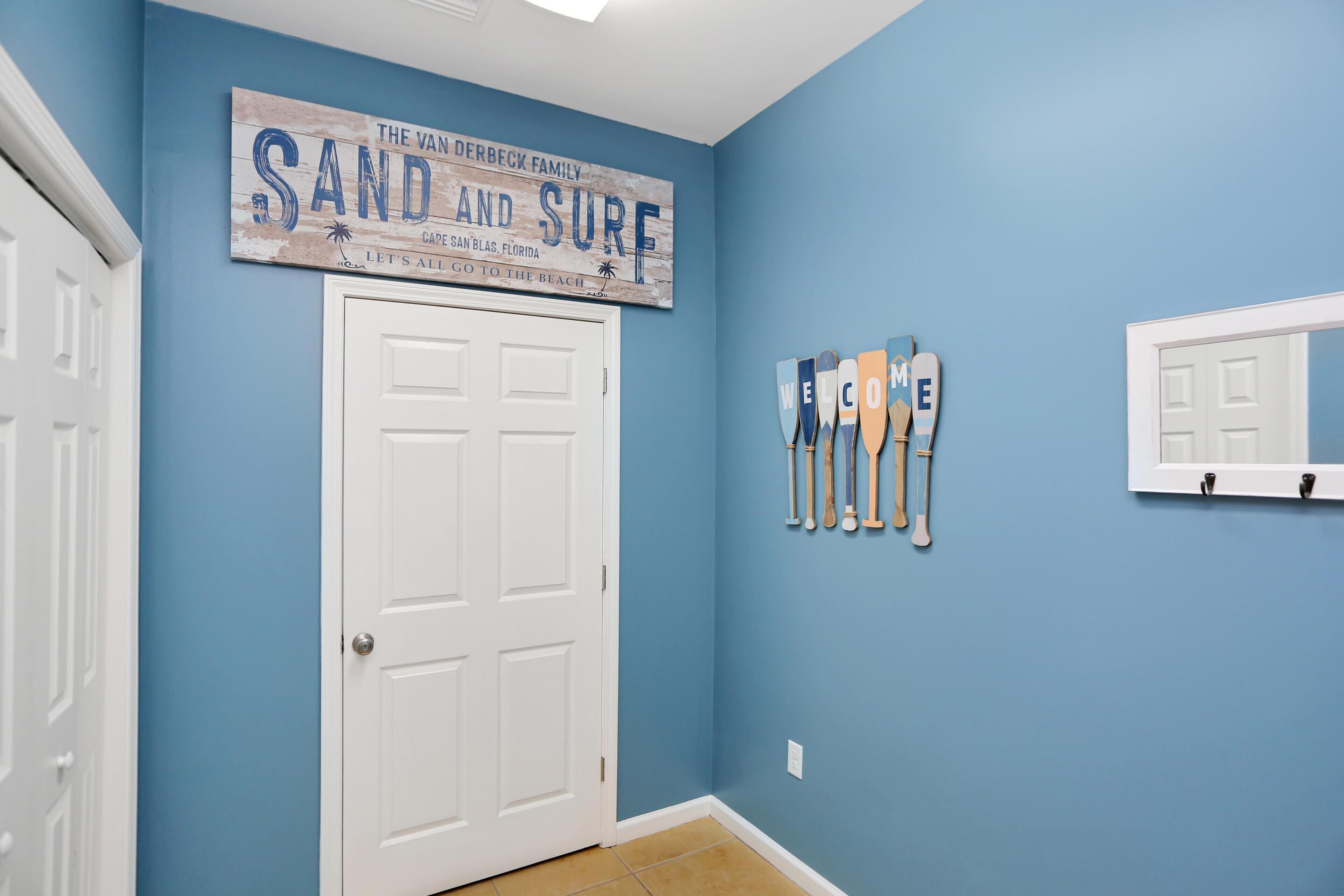 A hallway with a blue wall, a white door, and a sign that reads "THE VAN DERBECK FAMILY SAND AND SURF CAPE SAN BLAS, FLORIDA LET'S ALL GO TO THE BEACH."