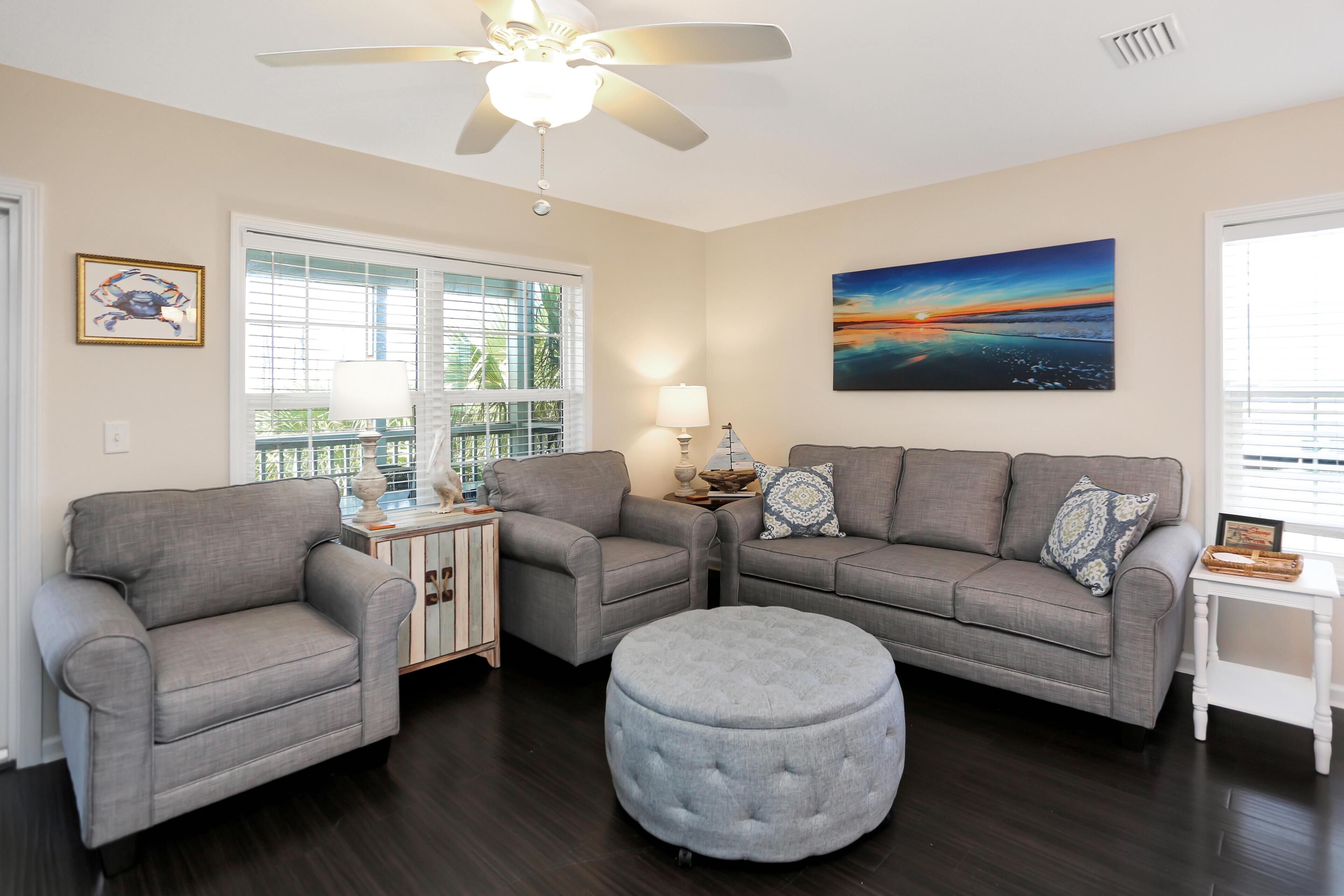 A living room with two grey sofas, a grey ottoman, a ceiling fan, and a painting of a sunset.
