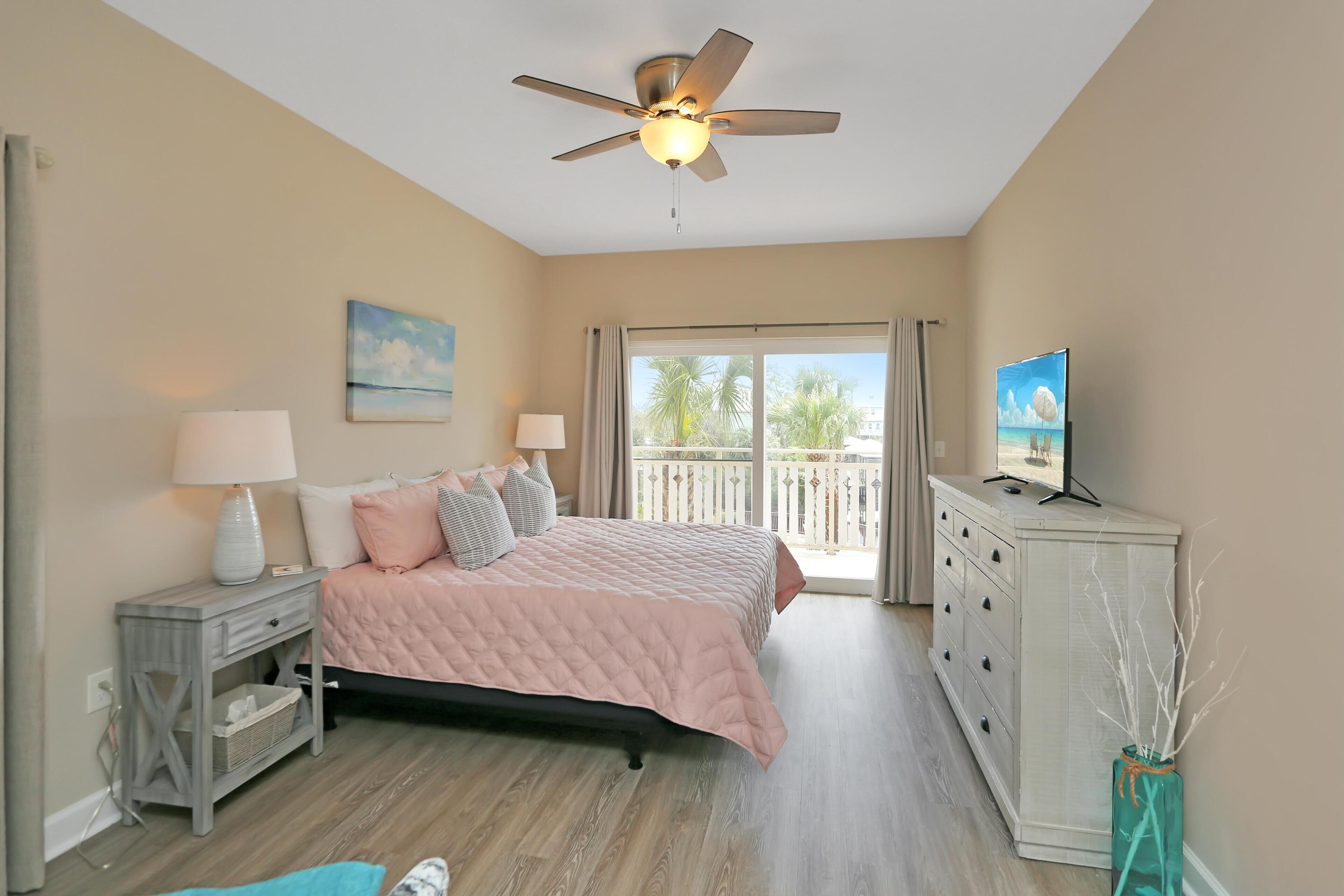 A bedroom with a bed, nightstand, dresser, TV, ceiling fan, and a balcony with a view.
