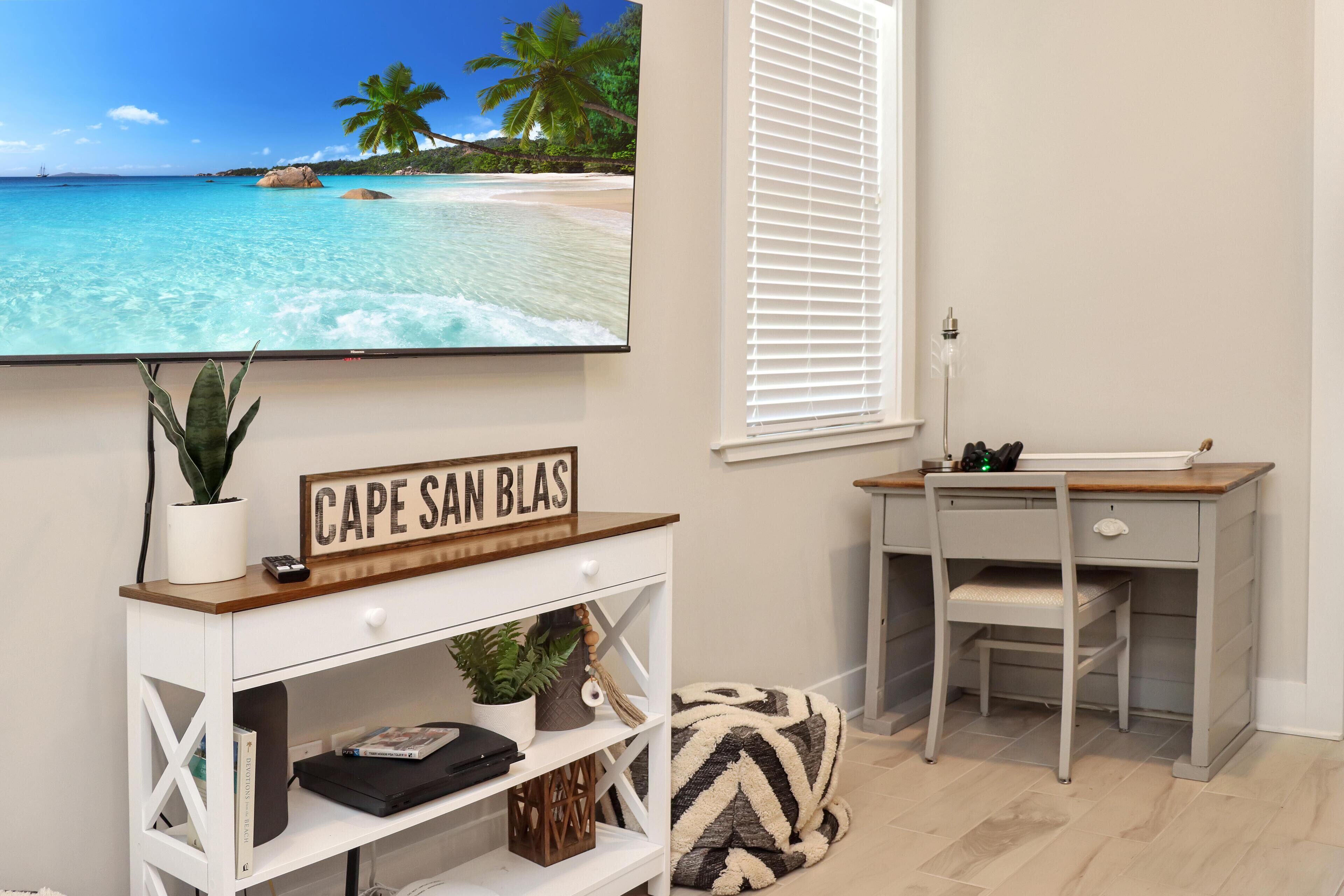 Ein Zimmer mit Fernseher, der eine Strandansicht zeigt, einem Schreibtisch mit Stuhl und einem Schild mit der Aufschrift 'CAPE SAN BLAS'.