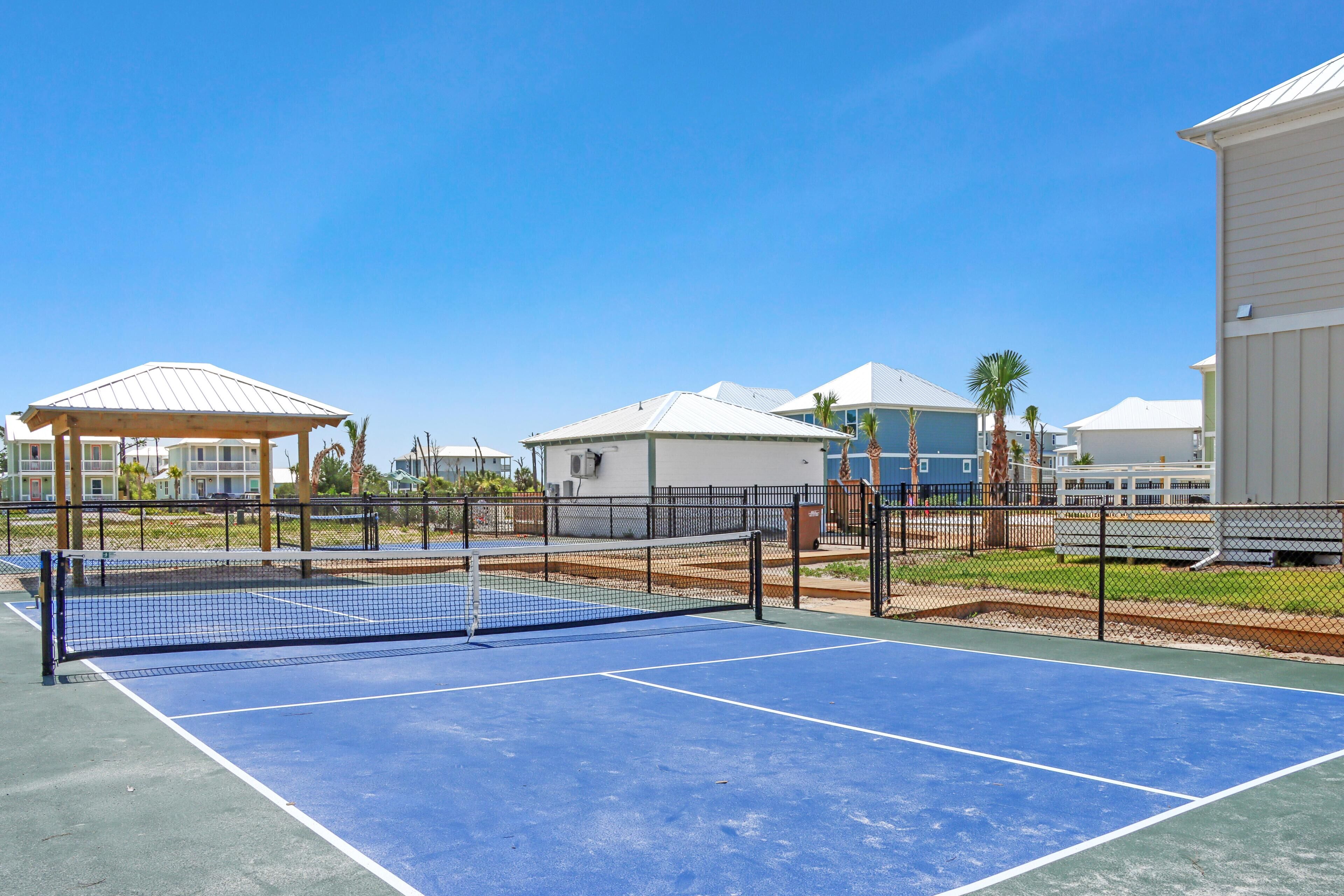 A tennis court with a net, surrounded by a fence, and adjacent to buildings with a white roof and a covered pavilion.