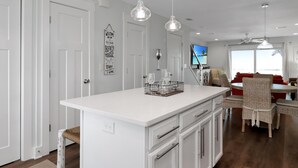 A modern kitchen with white cabinetry, a central island, pendant lighting, and a dining area with wicker chairs and a red sofa. - Wine Down (Port St. Joe)