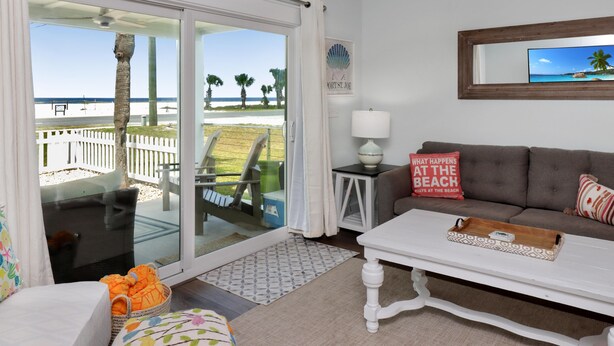 A beach house interior with a sliding glass door leading to a balcony, a white sofa, a small table, and a framed picture of a beach scene. - Wine Down (Port St. Joe)