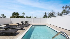 Una piscina rettangolare con acqua cristallina e di colore blu, circondata da una recinzione bianca. Sono presenti lettini e un tavolino con sedie intorno alla piscina.