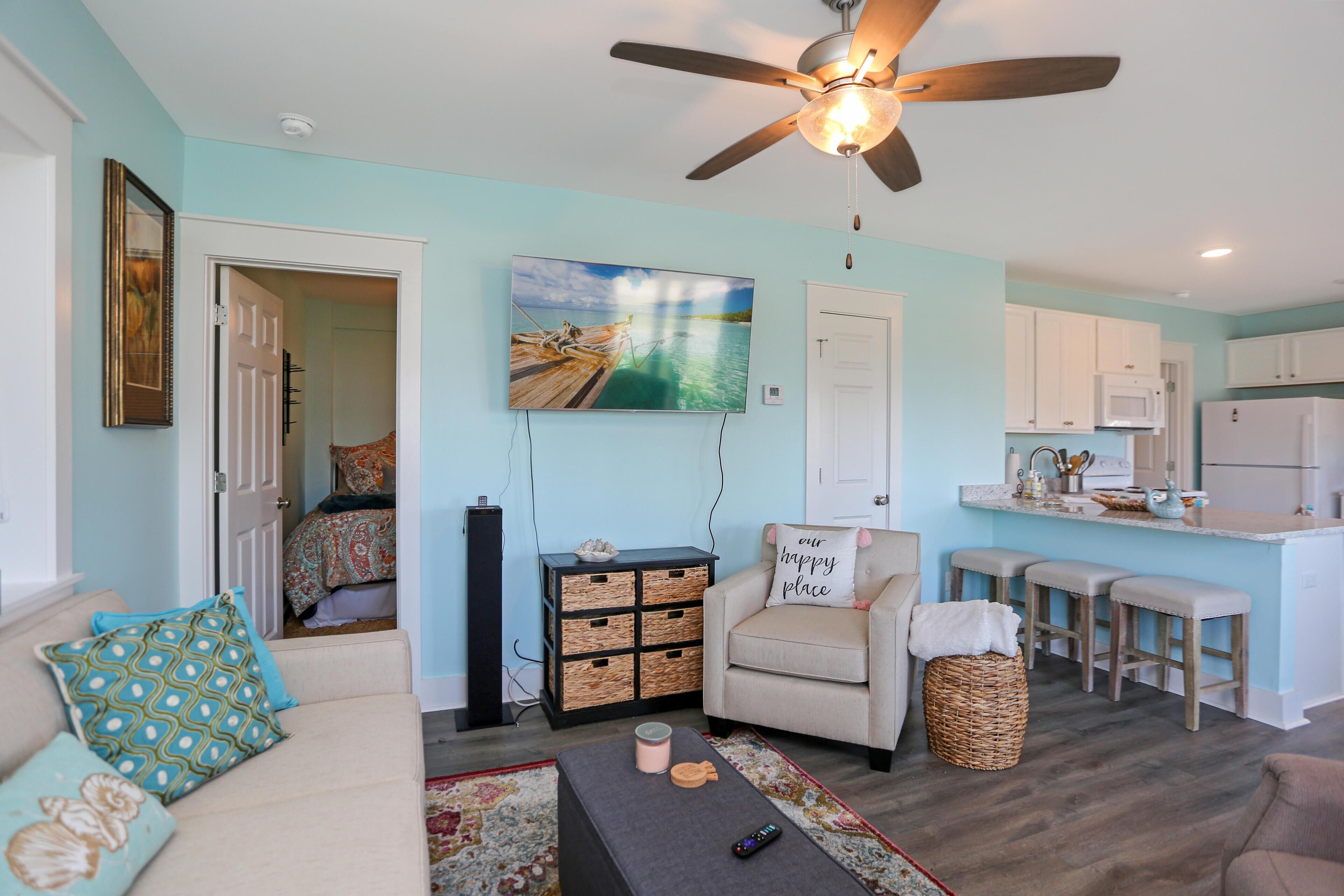 A living room with a sofa, a coffee table, a TV, a ceiling fan, and a kitchen area with a microwave and a refrigerator.