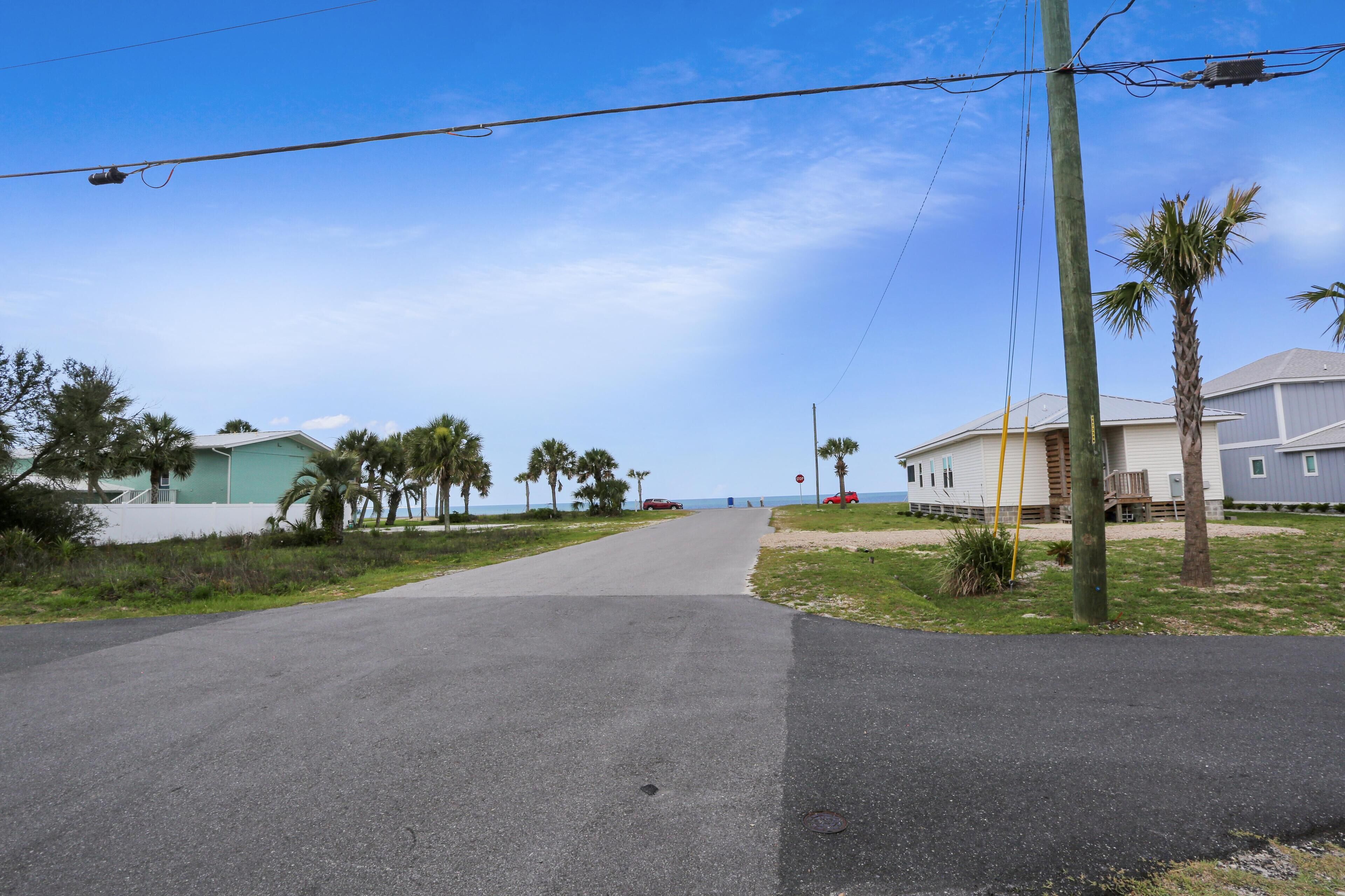 A paved road leading to a beachside community with single-story houses, palm trees, and a clear blue sky.