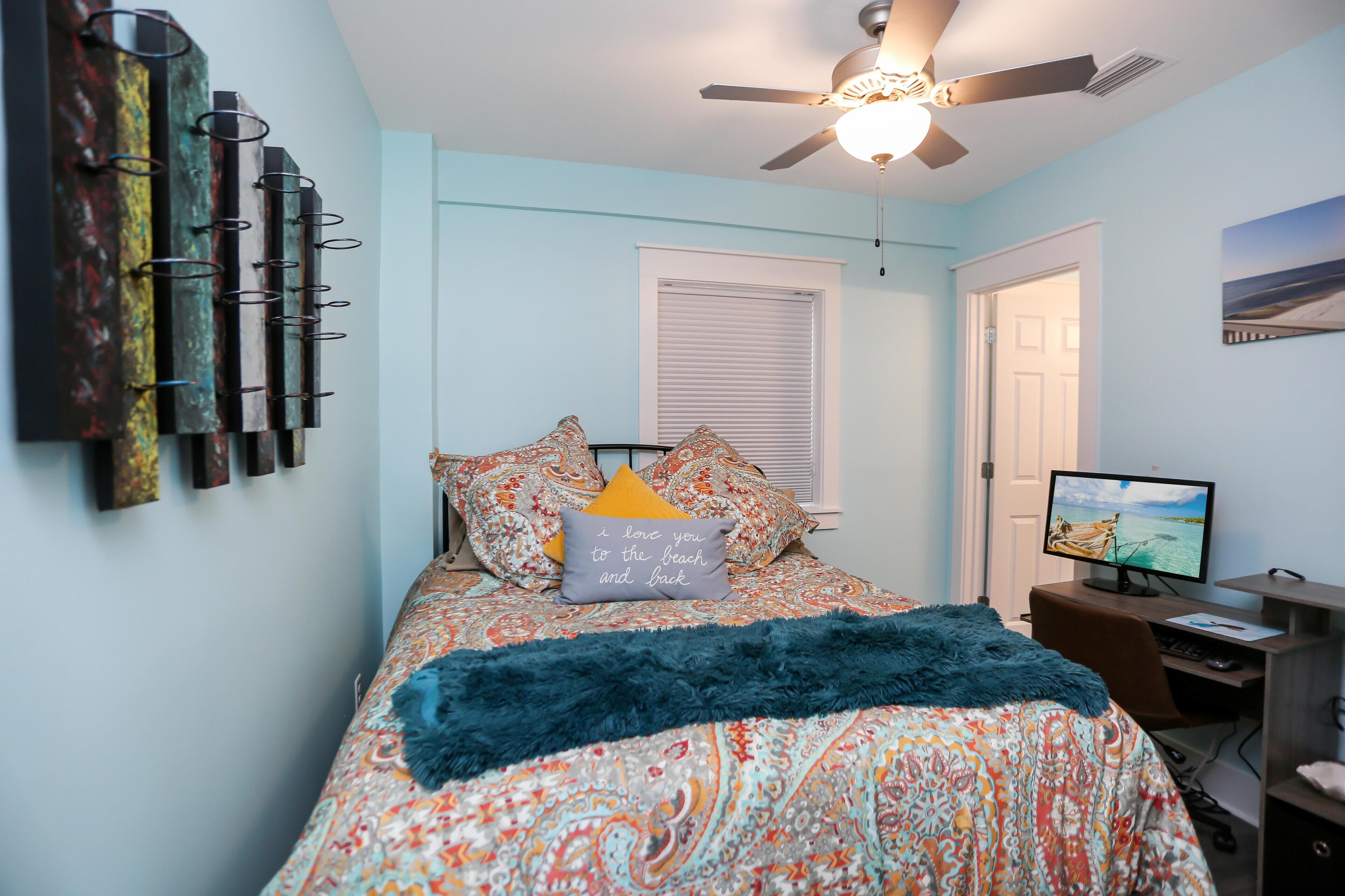 A bedroom with a bed, a desk, a computer, and a ceiling fan.