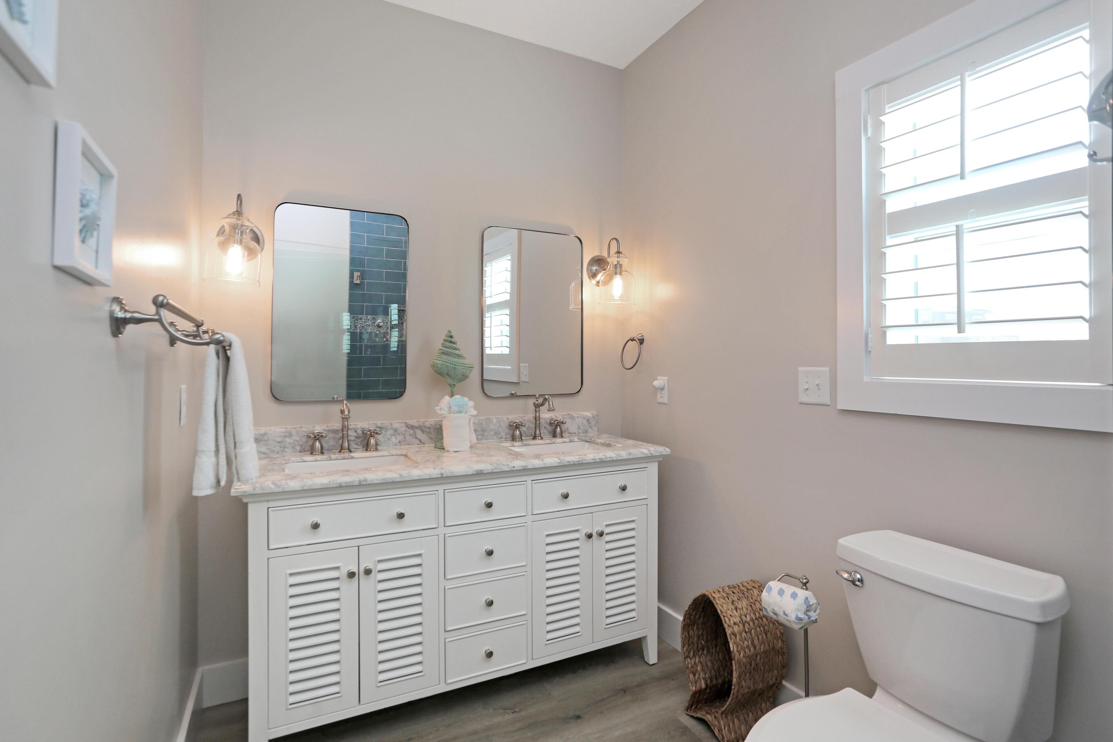A bathroom with a white vanity, two mirrors, a toilet, and a window with blinds.