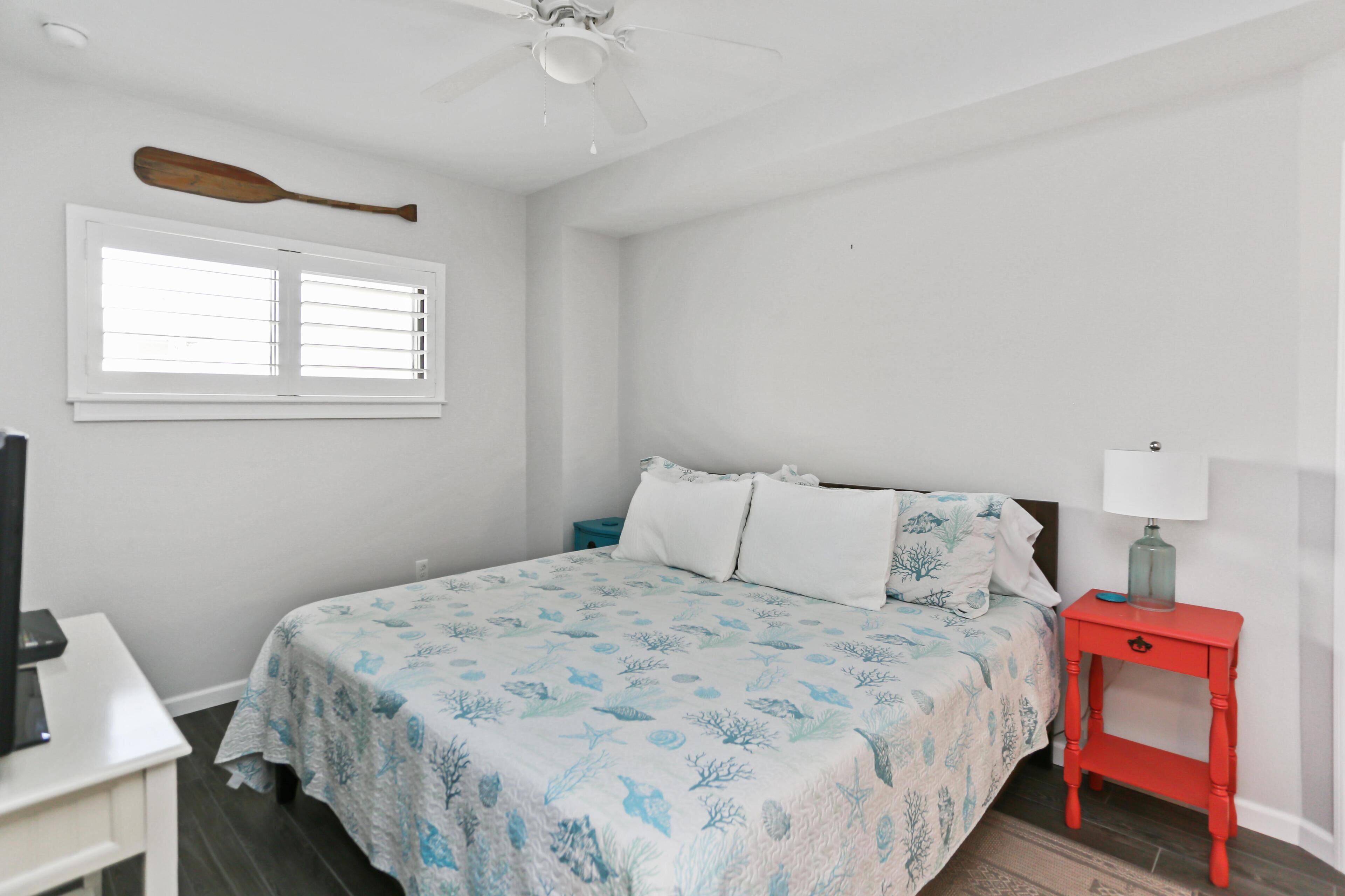 A bedroom with a bed, a nightstand, a ceiling fan, a window, and a wall-mounted paddleboard.