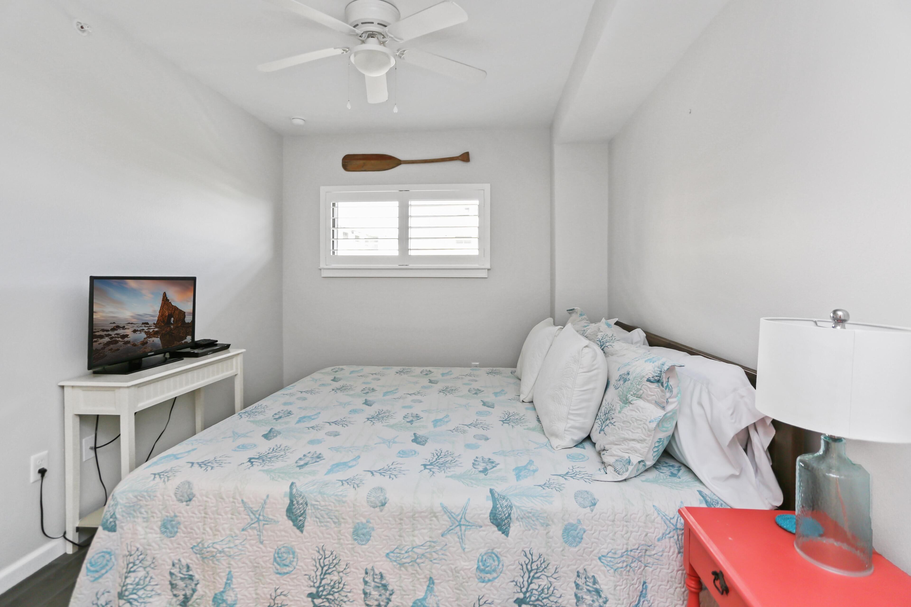 Un dormitorio con una cama, un televisor sobre un soporte, una ventana y un ventilador de techo.