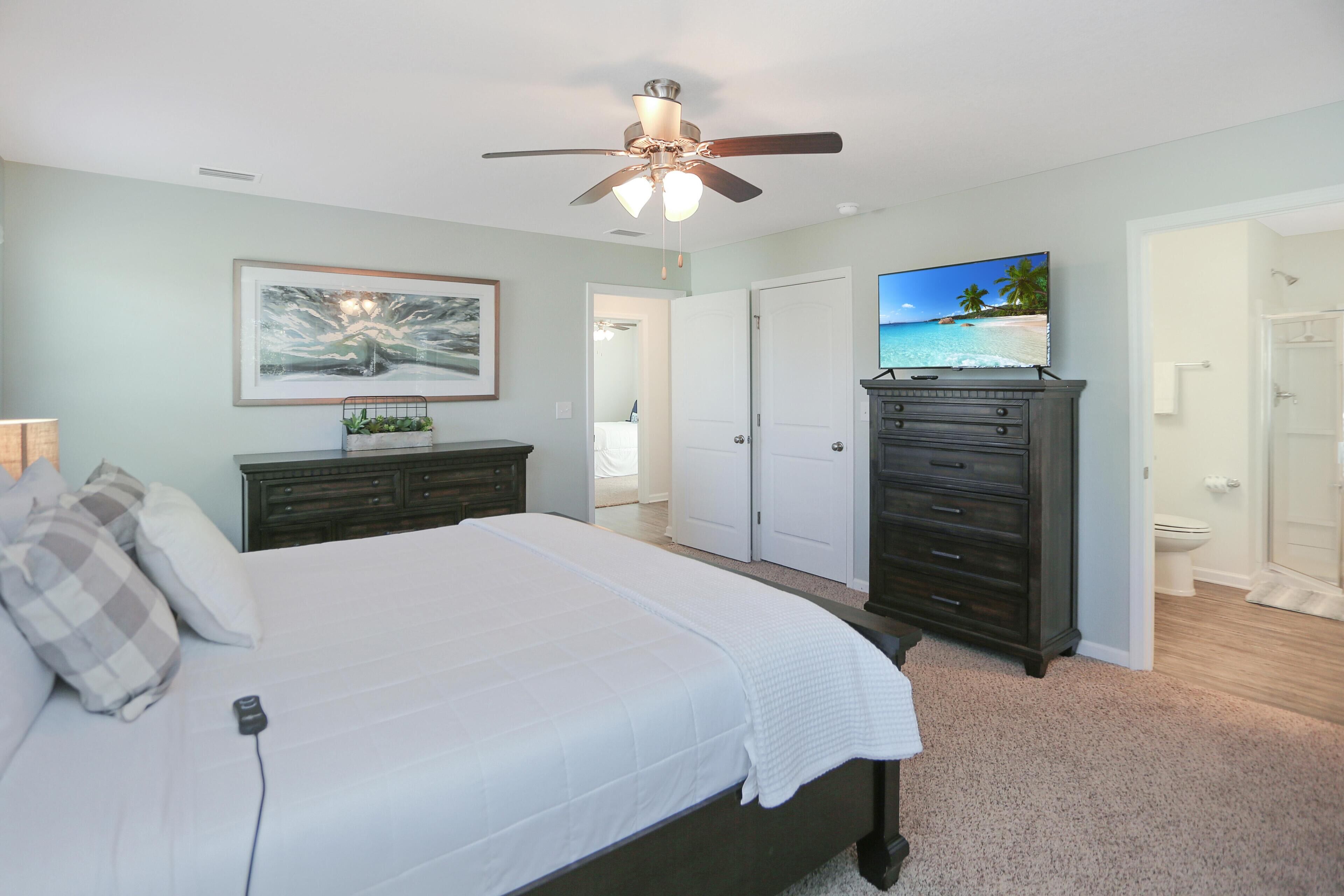 A bedroom with a bed, a dresser, a ceiling fan, a TV, and a bathroom with a shower and toilet.