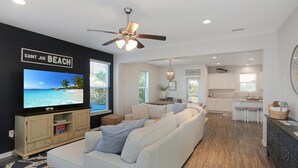 A modern living room with a large flat-screen TV, a ceiling fan, and a kitchen area in the background. - Song of the Sea (Port St. Joe)