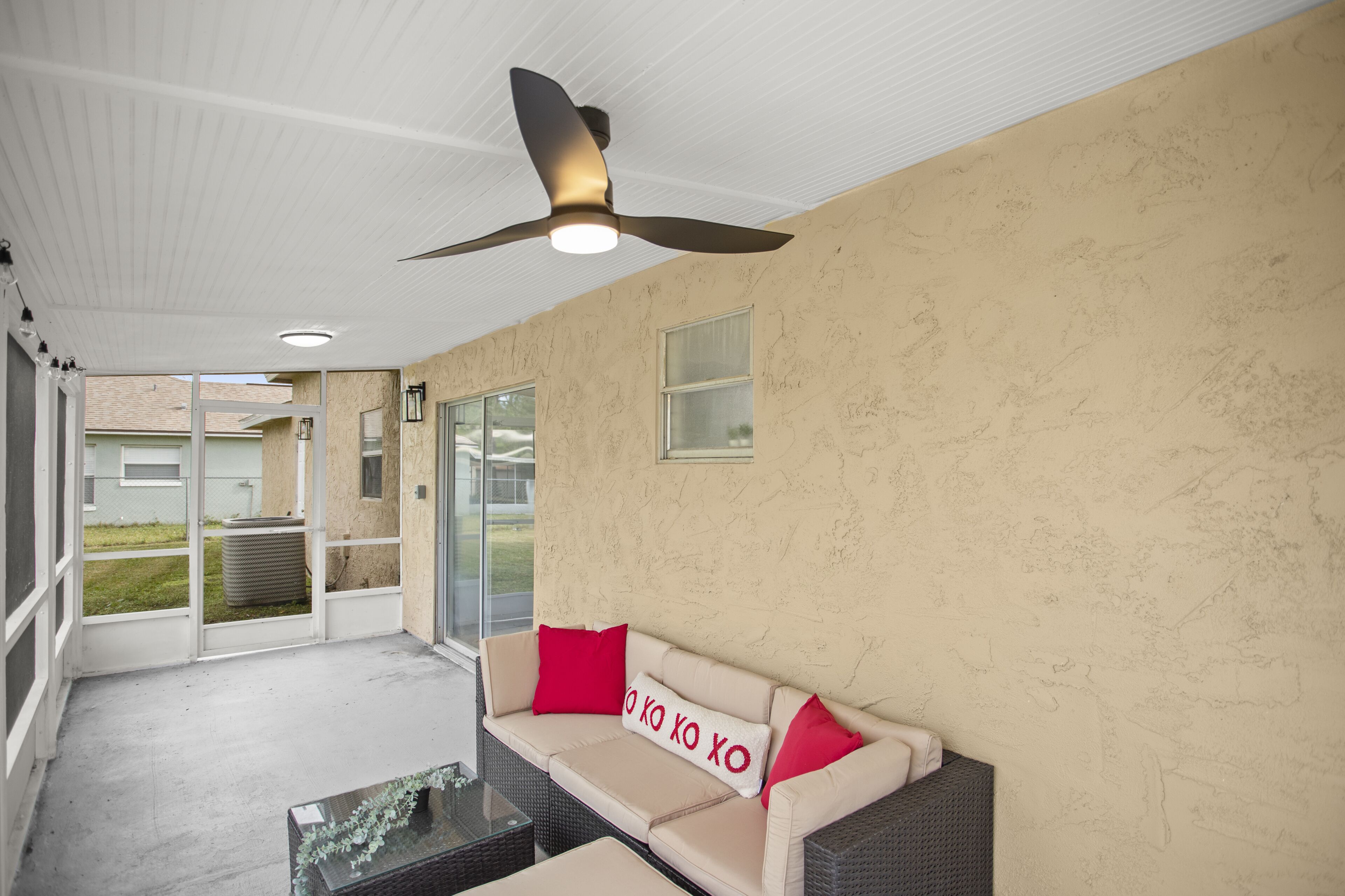 Un patio cubierto con una pared color beige, un ventilador de techo, una puerta de vidrio que da al exterior y un sofá color beige con cojines rojos.