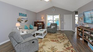 A living room with a ceiling fan, a couch, a chair, a coffee table, and a bookshelf. - The Lighthouse (Port St. Joe)