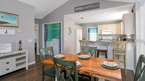 A dining area with a wooden table and green chairs, a kitchen with white cabinets, and a sign on the wall that reads "Happiness is a day at the beach." - The Lighthouse (Port St. Joe)