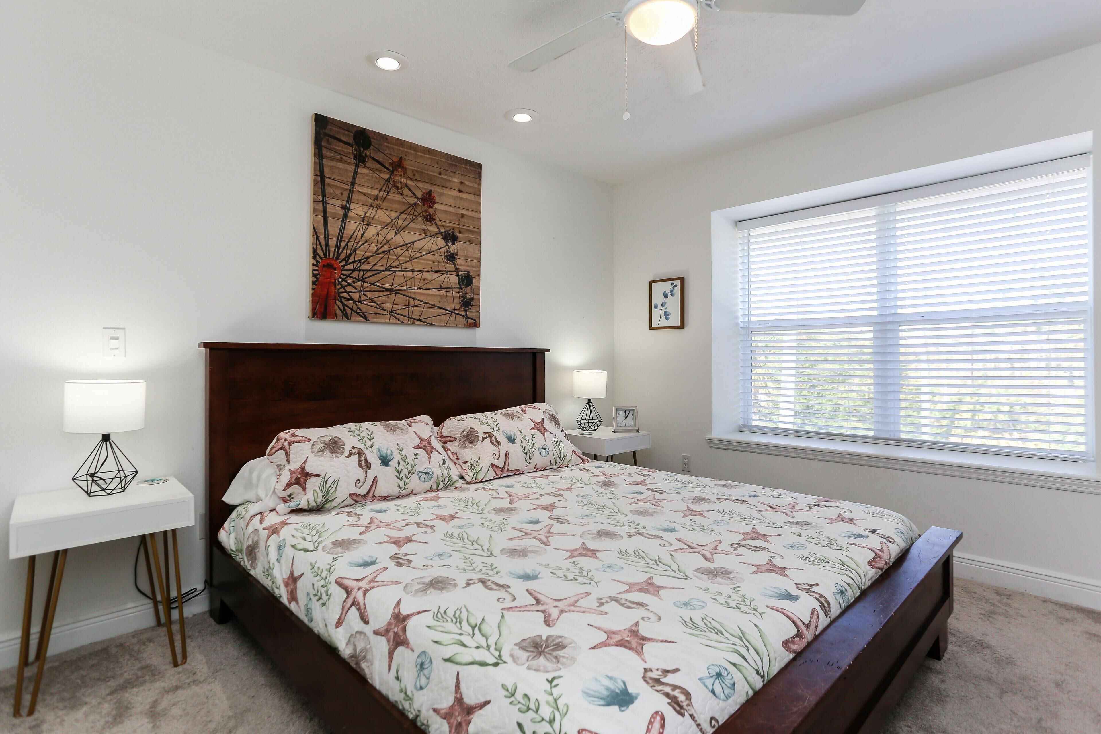 A bedroom with a large bed, a ceiling fan, a window with blinds, and a wall art piece.
