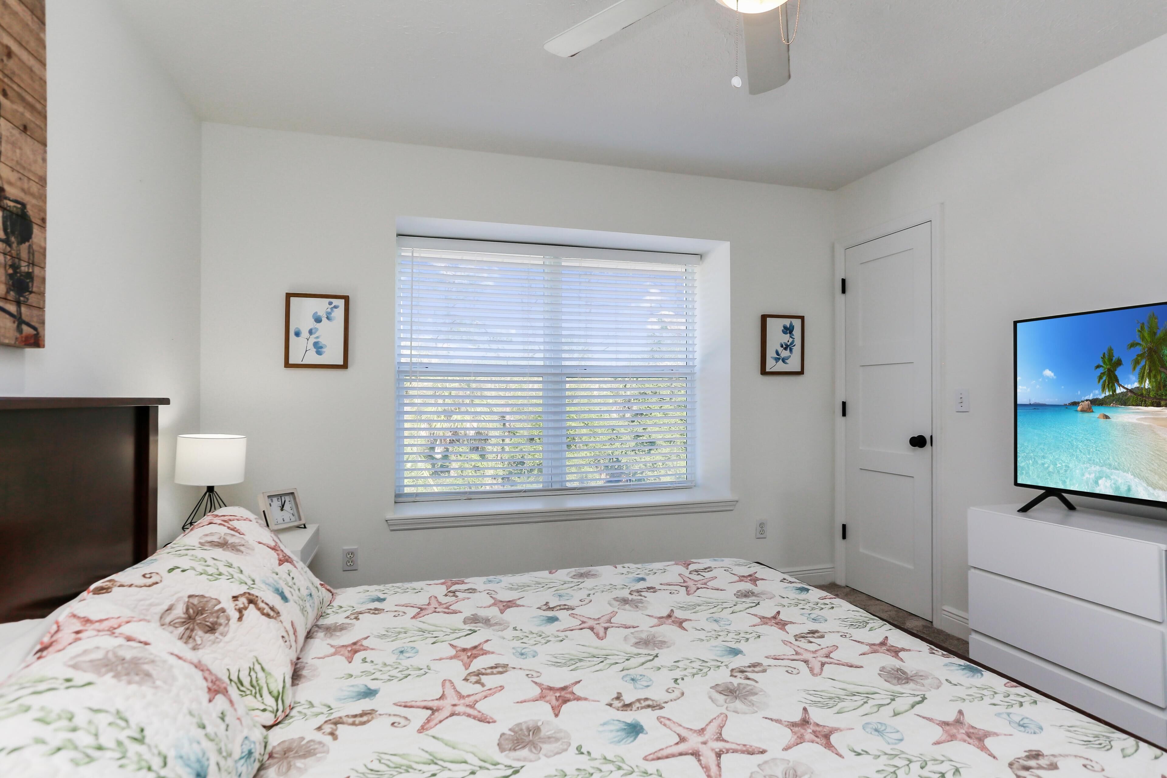 Un dormitorio con cama, televisión, ventana con persianas y puerta.