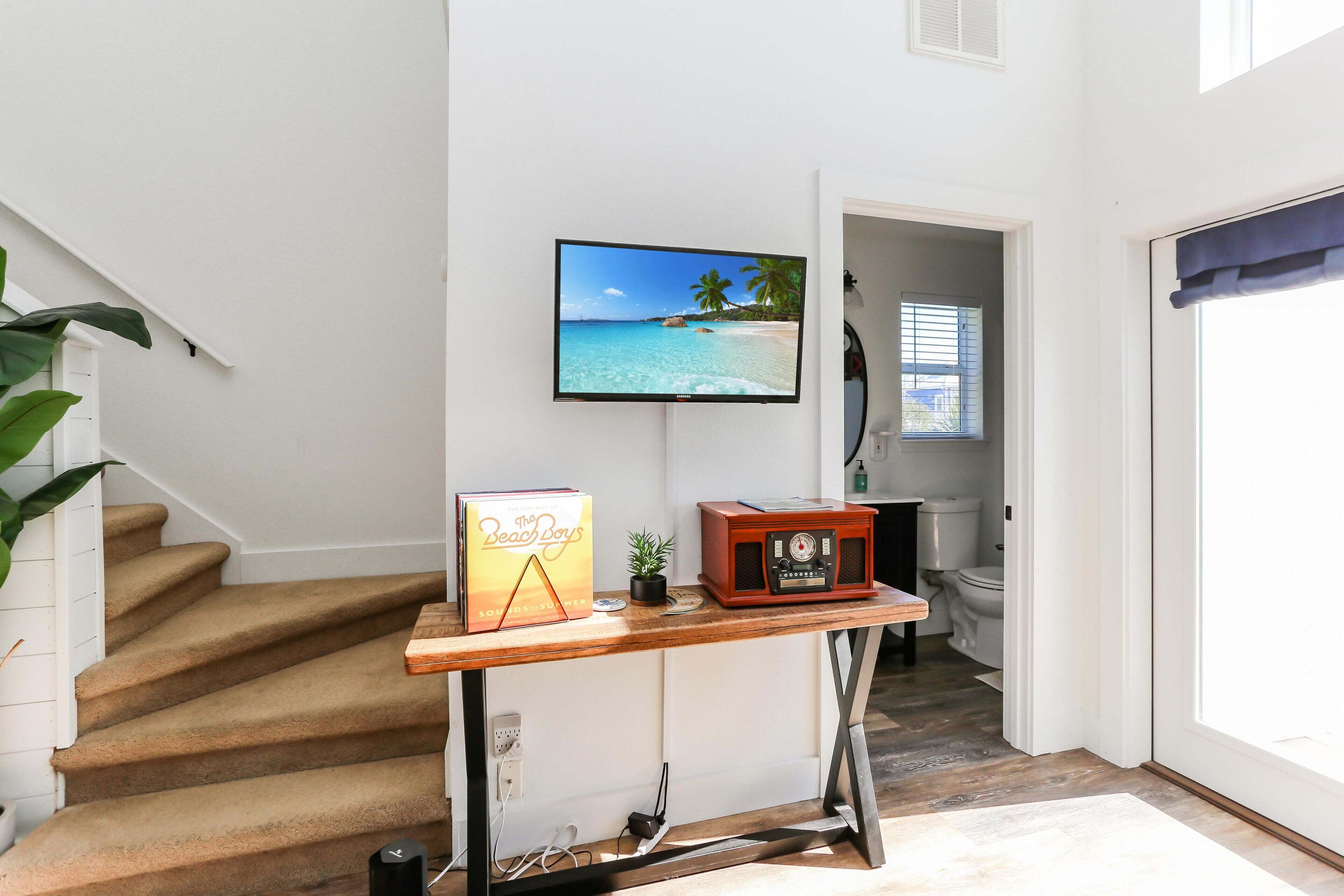 A staircase with a wooden table featuring a book, a small plant, and a vintage radio. A TV on the wall displays a beach scene.