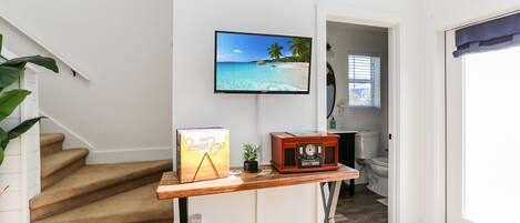 A staircase with a wooden table featuring a book, a small plant, and a vintage radio. A TV on the wall displays a beach scene.