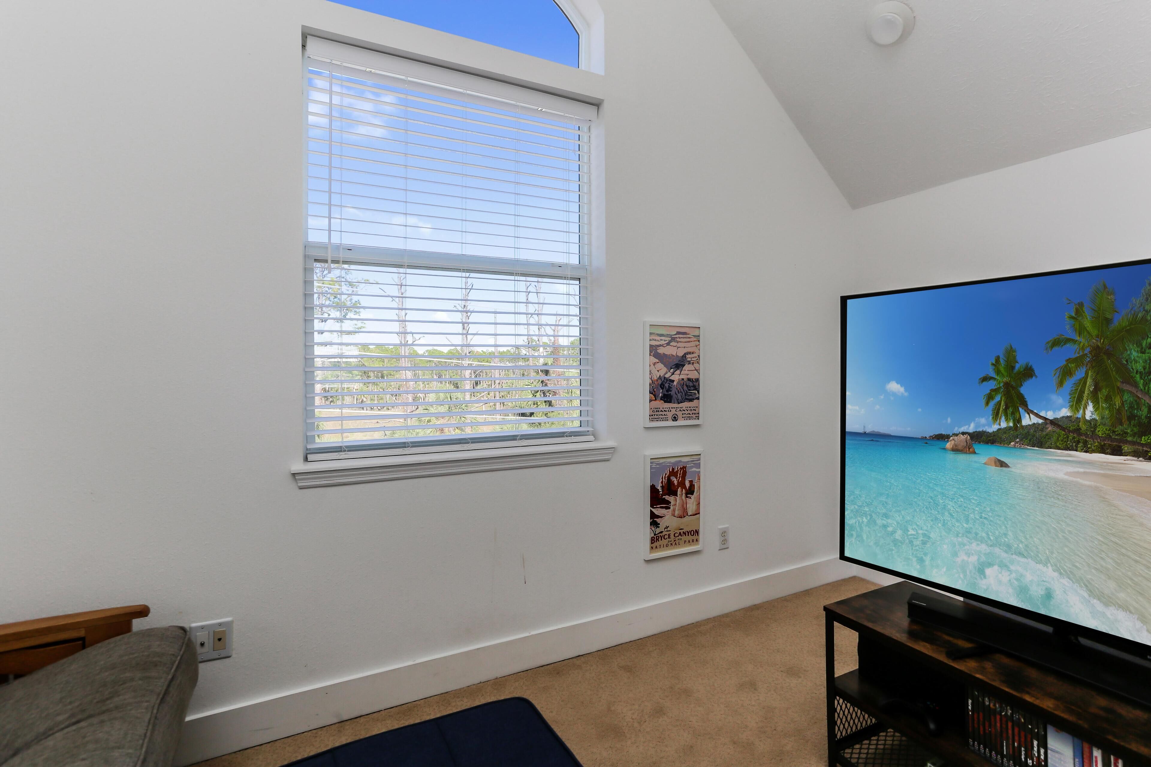 A living room with a large flat-screen TV, a window with blinds, a couch, and a small bookshelf.