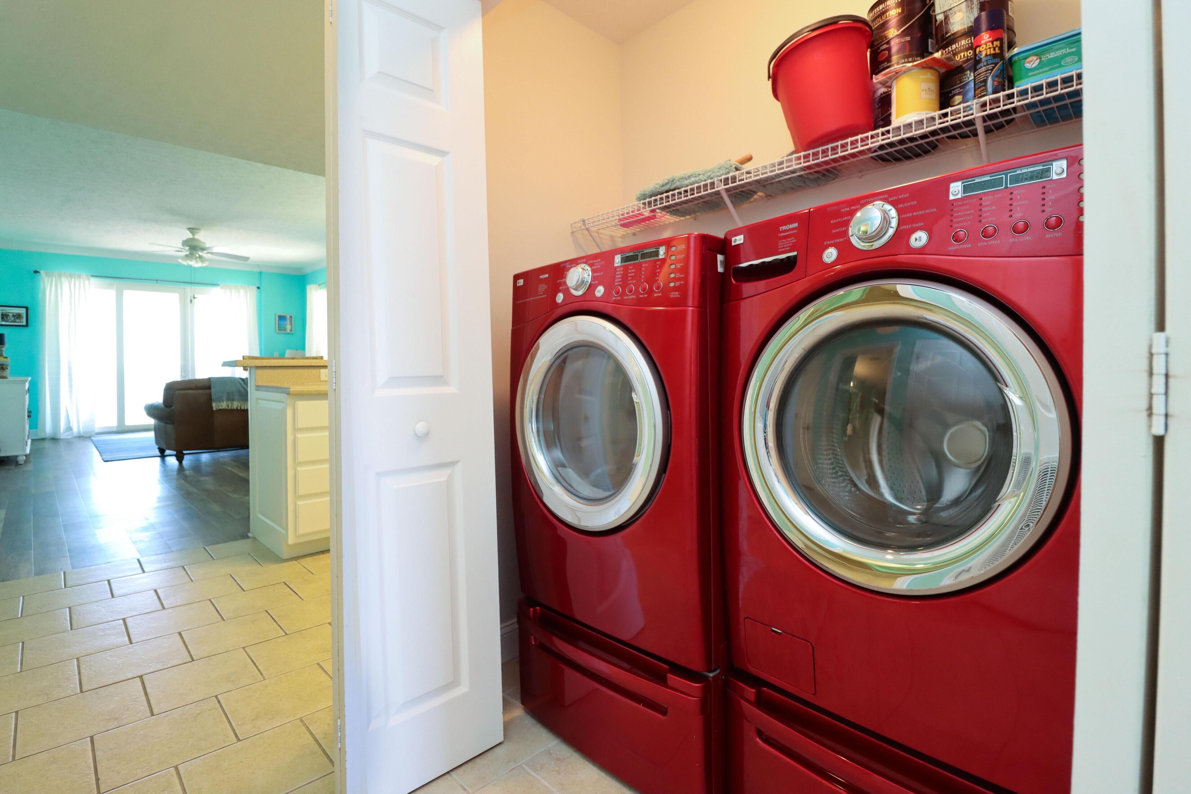 Une buanderie avec deux machines à laver et deux sèche-linge à chargement frontal de couleur rouge, une porte blanche et une étagère au-dessus des sèche-linge où sont rangés divers objets.