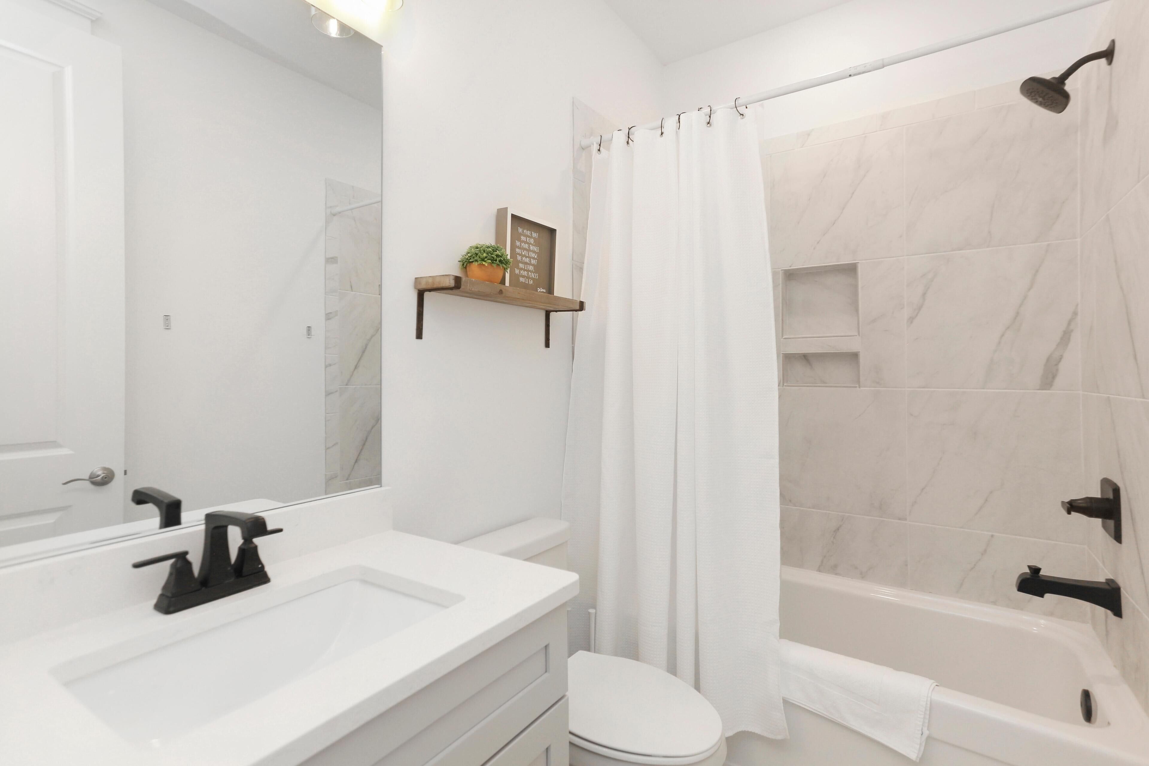 A modern bathroom with a white sink, black faucet, and a white toilet. There is a shower curtain and a shelf with a plant and a framed quote.