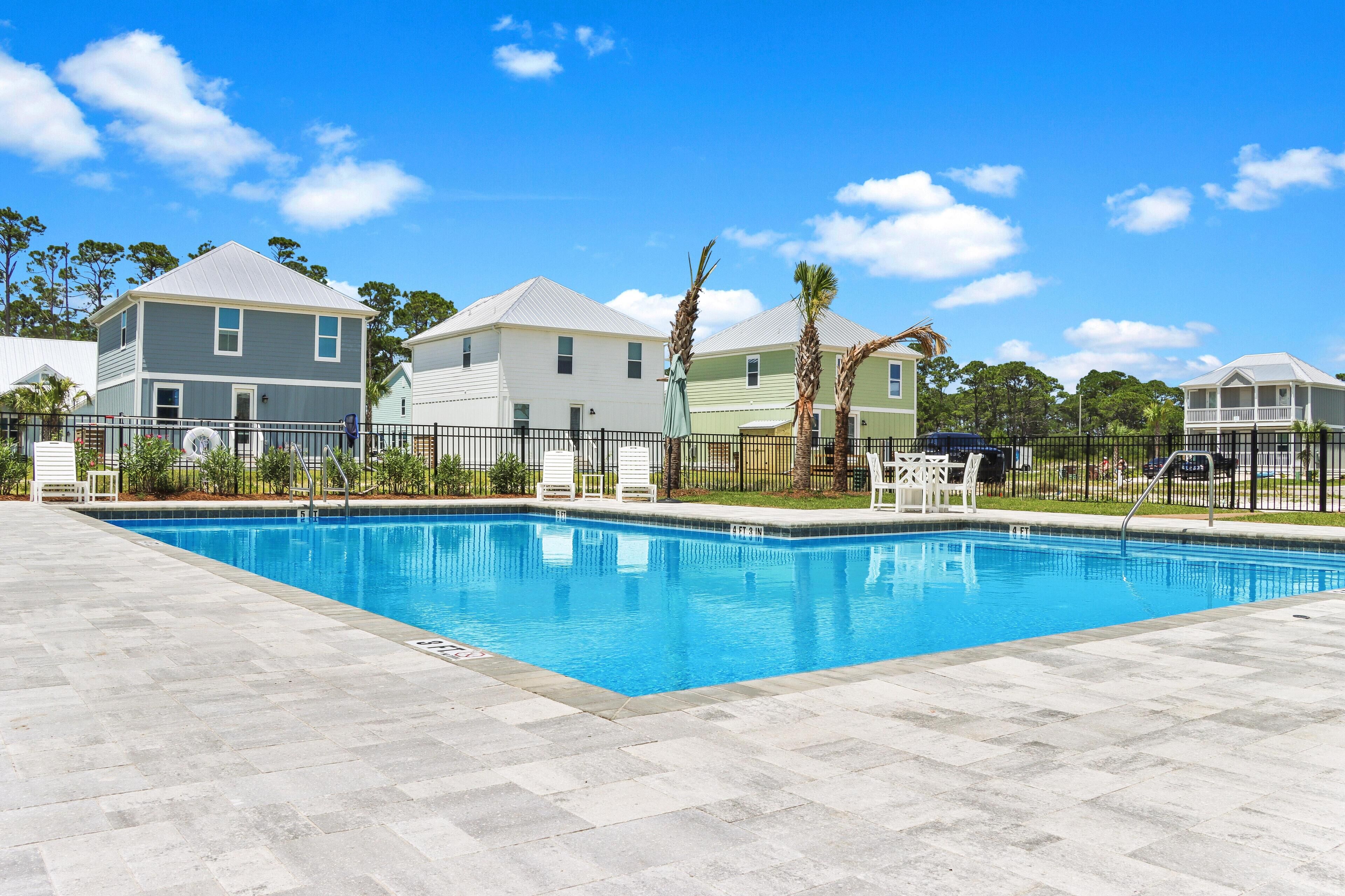 Una piscina con suelo de baldosas, rodeada por una valla, y casas al fondo.