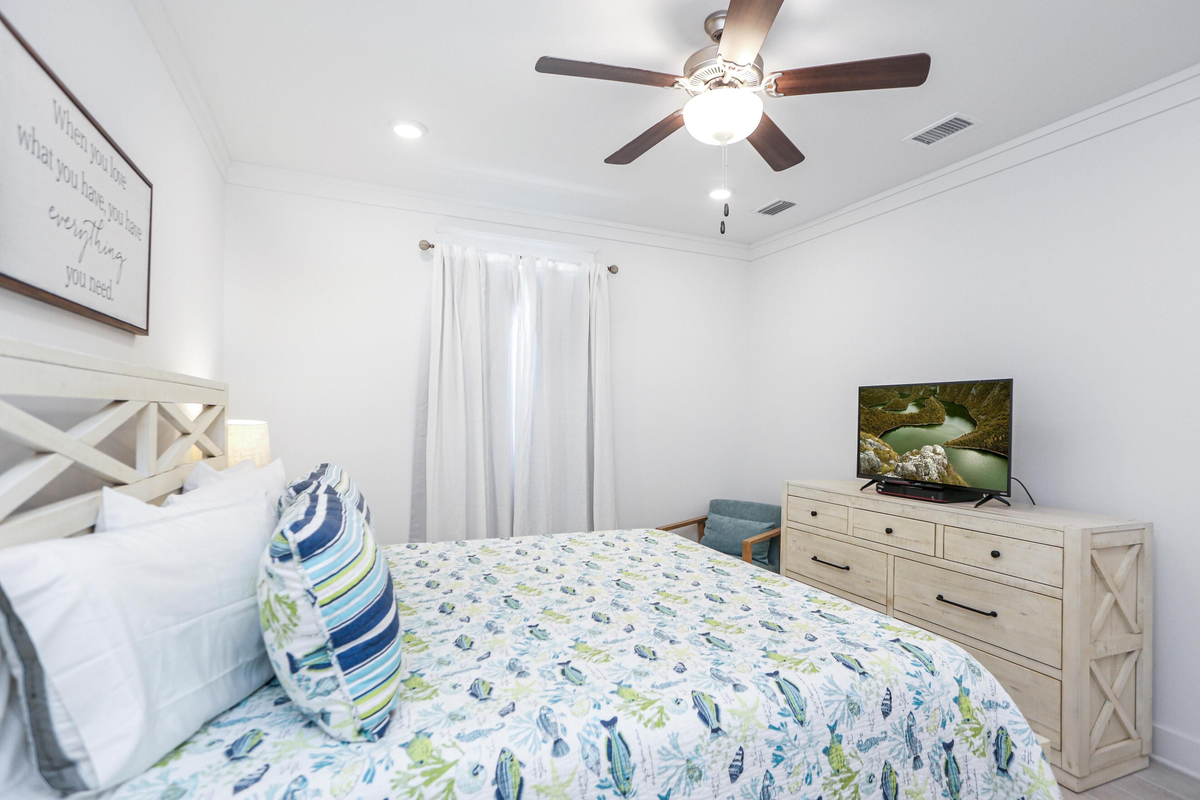 A bedroom with a bed, a ceiling fan, a TV, a dresser, and a window with curtains.