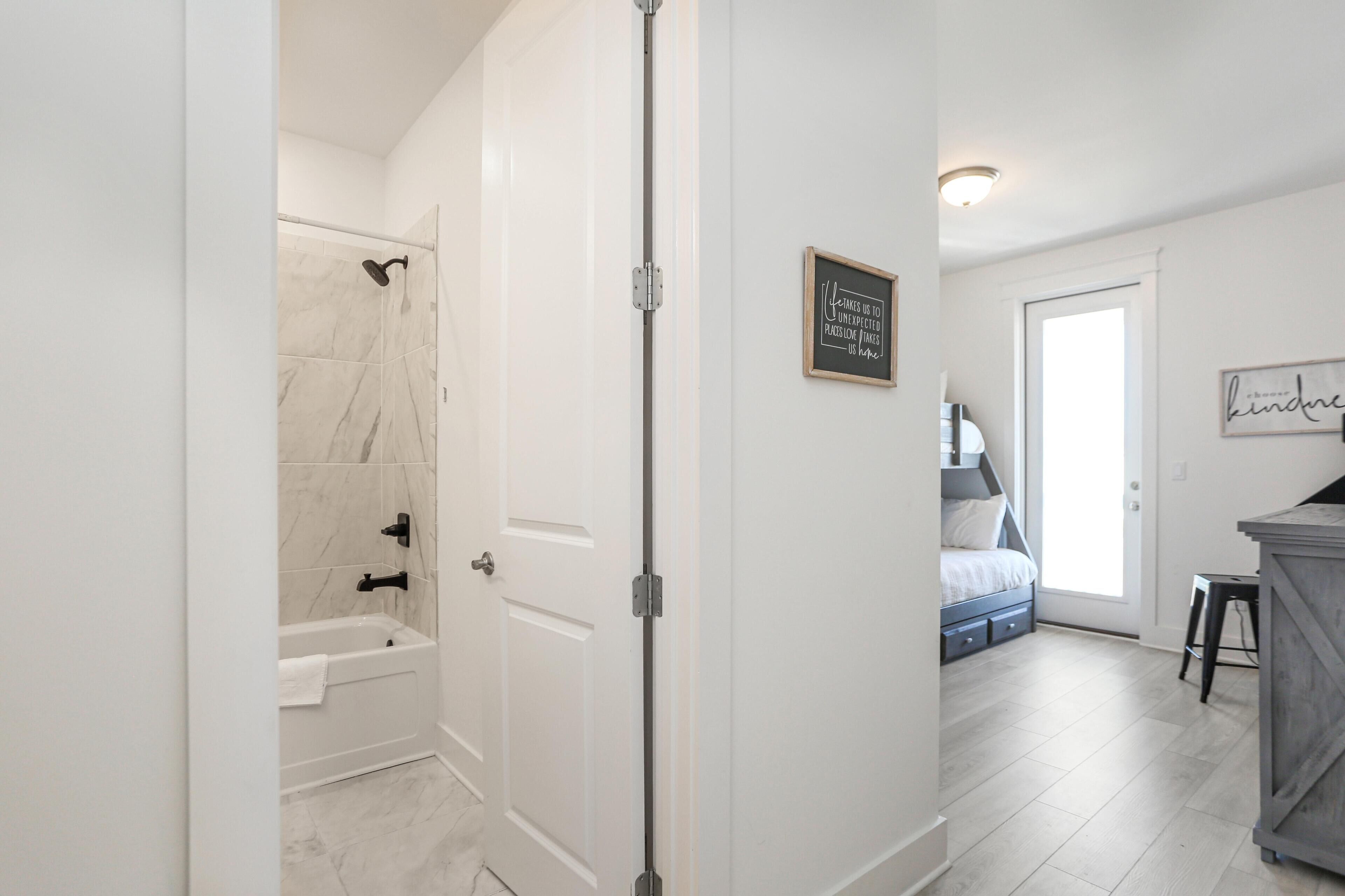 Een badkamer met een douche, een bad en een toilet. Een deur leidt naar een slaapkamer met een bed en een dressoir.