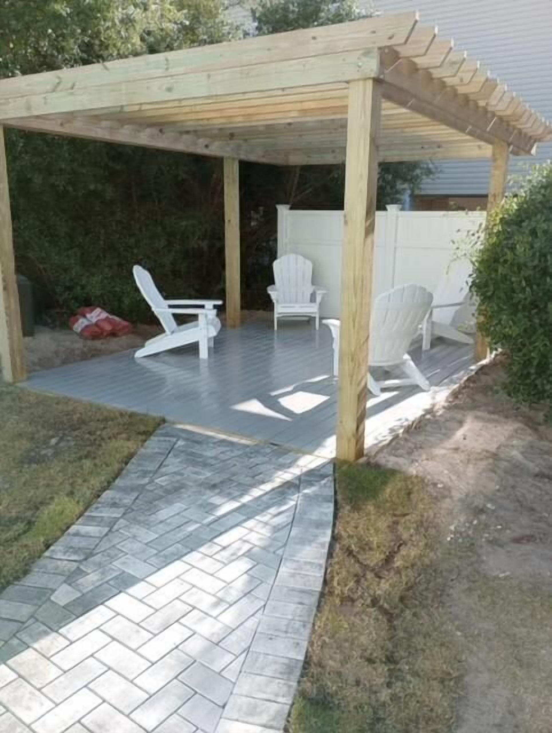 Un espacio de patio cubierto con una pérgola de madera, dos sillas Adirondack blancas y un sendero empedrado.