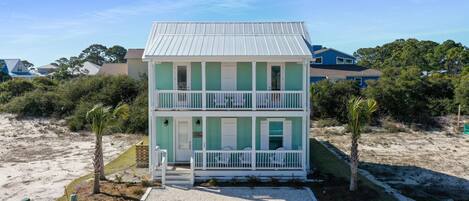 A two-story beach house with a white exterior and green accents, featuring a front porch and a small deck.