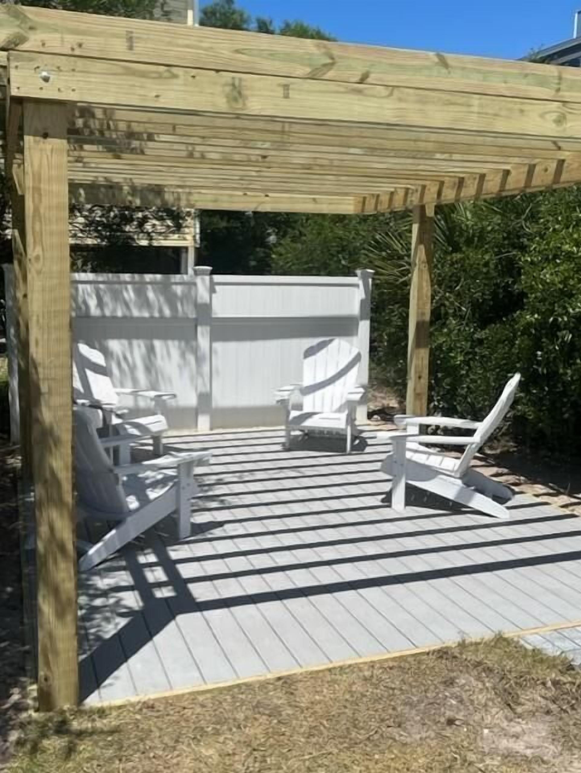 A wooden deck with two white lounge chairs and a small table, surrounded by a wooden pergola and a white fence.