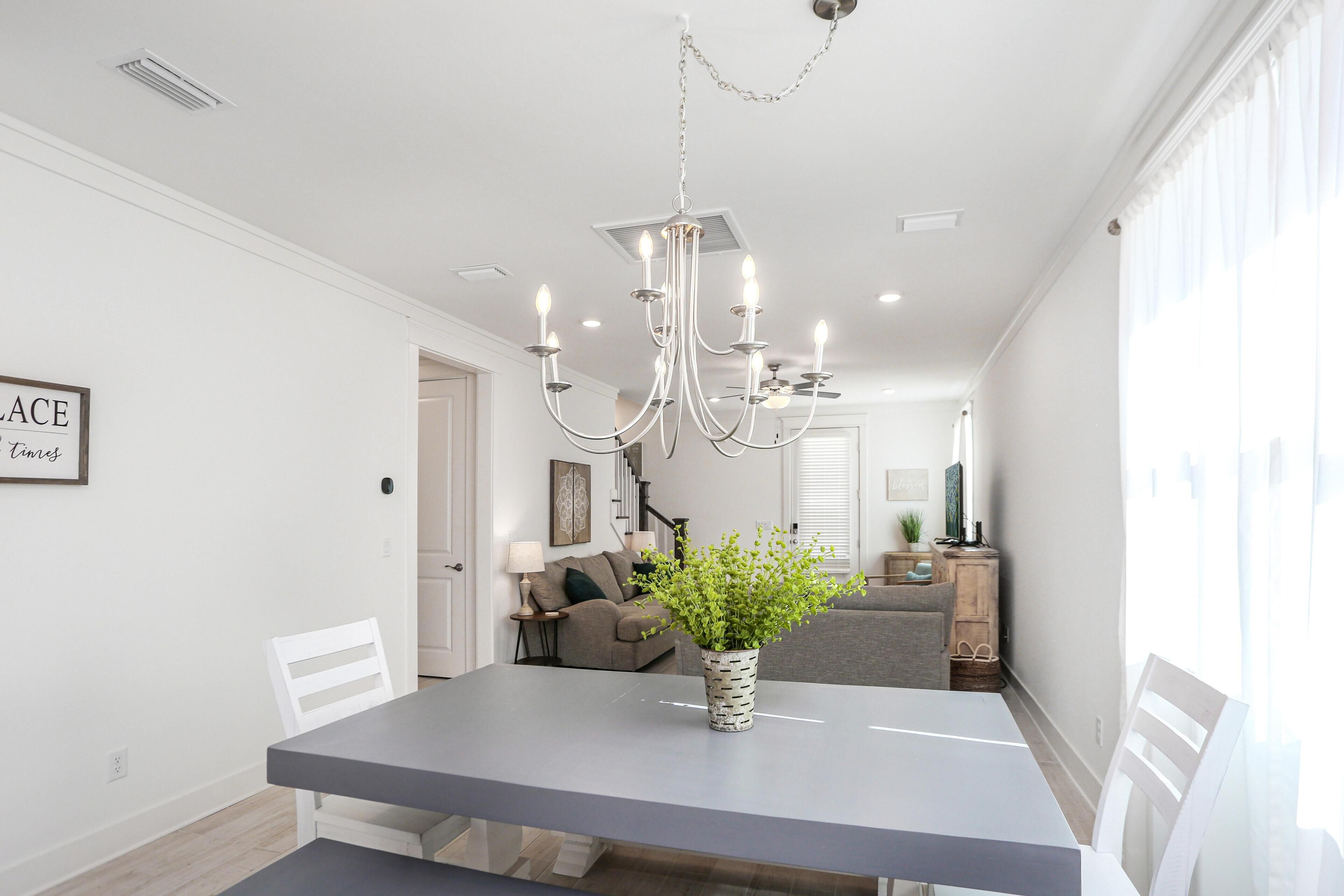A dining area with a grey table, white chairs, a chandelier, and a potted plant.