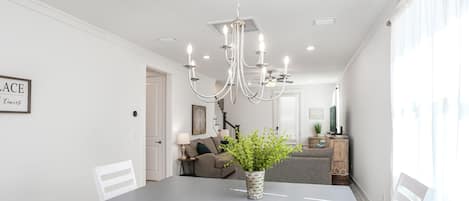 A dining area with a grey table, white chairs, a chandelier, and a potted plant.