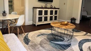 Un salon comprenant une table basse ronde, un téléviseur fixé au mur, un coin repas avec une table et des chaises blanches, et un meuble avec des portes en verre.