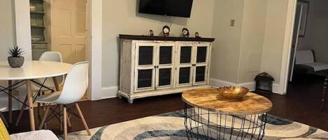 A living room with a round coffee table, a television mounted on the wall, a dining area with a white table and chairs, and a cabinet with glass doors.