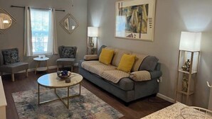 A living room with a grey sofa, a coffee table, and a ceiling fan. There are two windows with curtains, a lamp, and a framed picture on the wall.