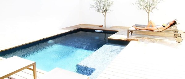 Une piscine avec une terrasse en bois, une chaise longue et un petit arbre.