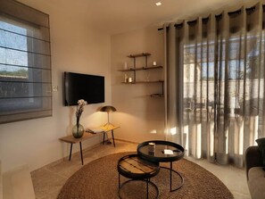 Uma sala de estar moderna com uma televisão de tela plana, uma mesinha com abajur, uma mesa de centro redonda e uma janela grande com cortinas.