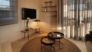 Ein modernes Wohnzimmer mit Fernseher, einem kleinen Tisch mit Lampe, einem runden Couchtisch und einem großen Fenster mit Vorhängen.